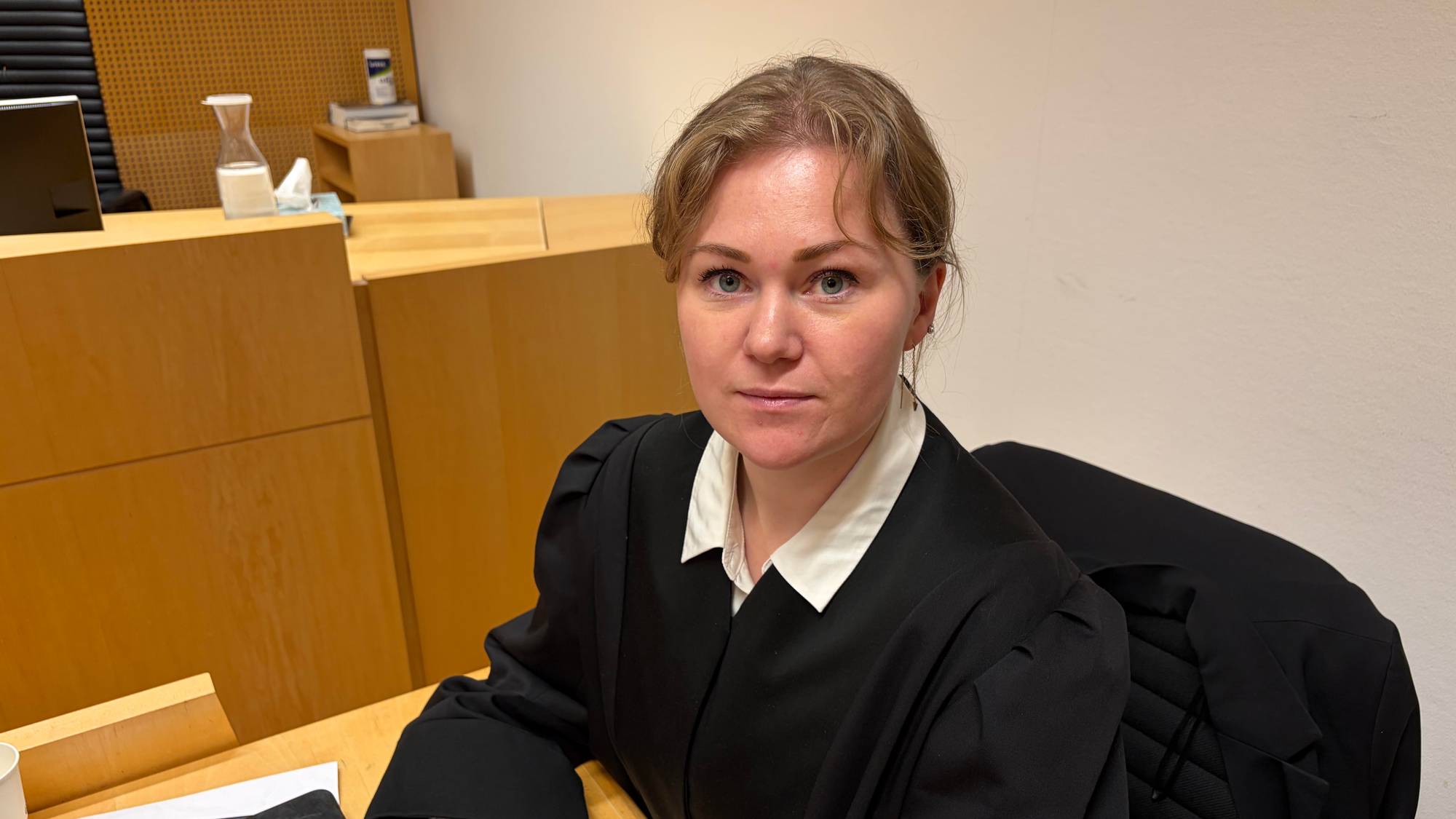 En kvinne med blondt hår ser inn i kamera. Hun har på seg en mørk kappe.