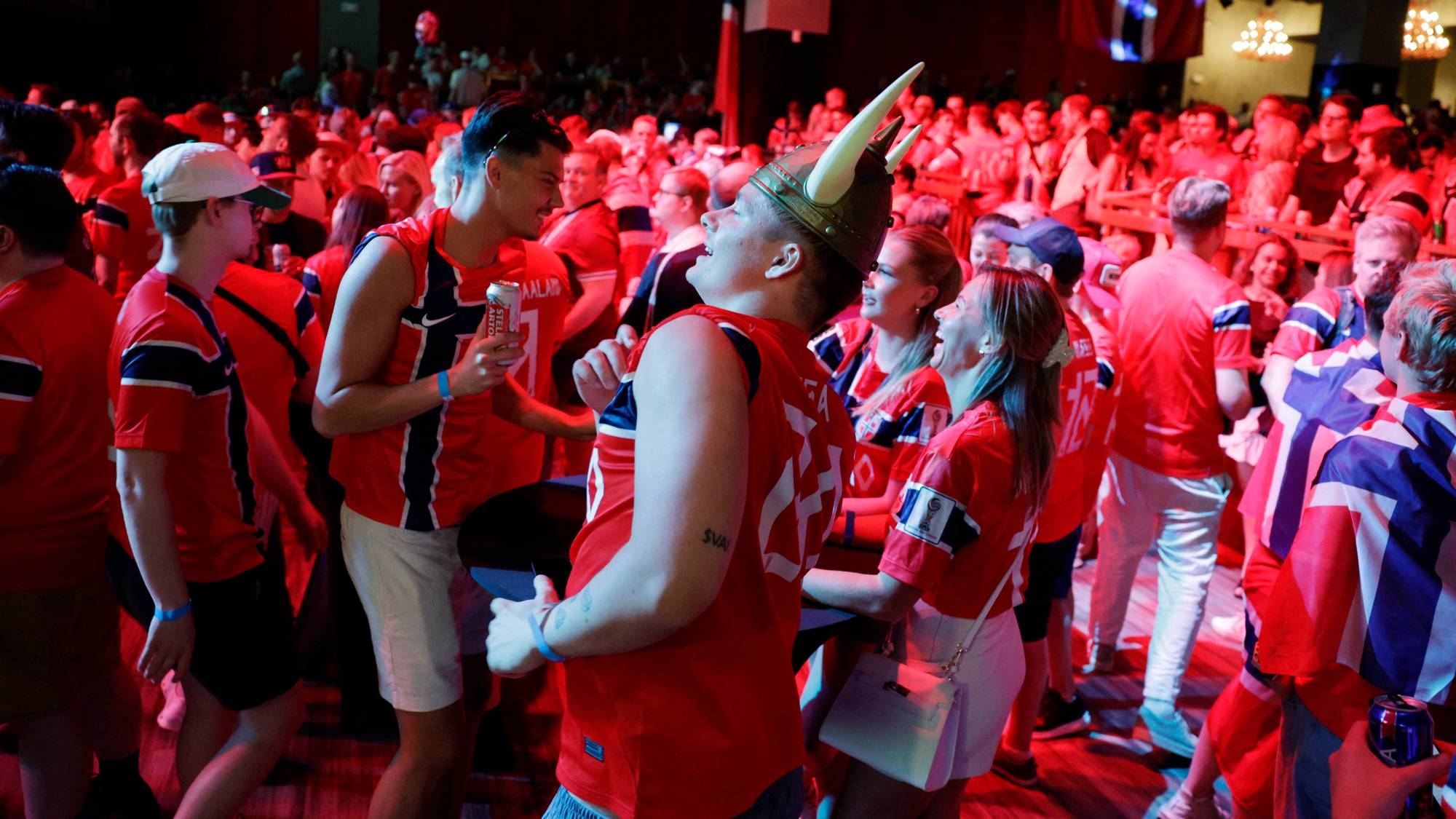 En gruppe mennesker, kledd i Norges supporterdrakter og flagg, samles innendørs. En person med vikinghjelm er sentral i bildet. Stemningen er livlig, med mange som smiler og holder drikke. Belysningen er rød og blå. (Bildebeskrivelsen er laget av en KI-tjeneste)