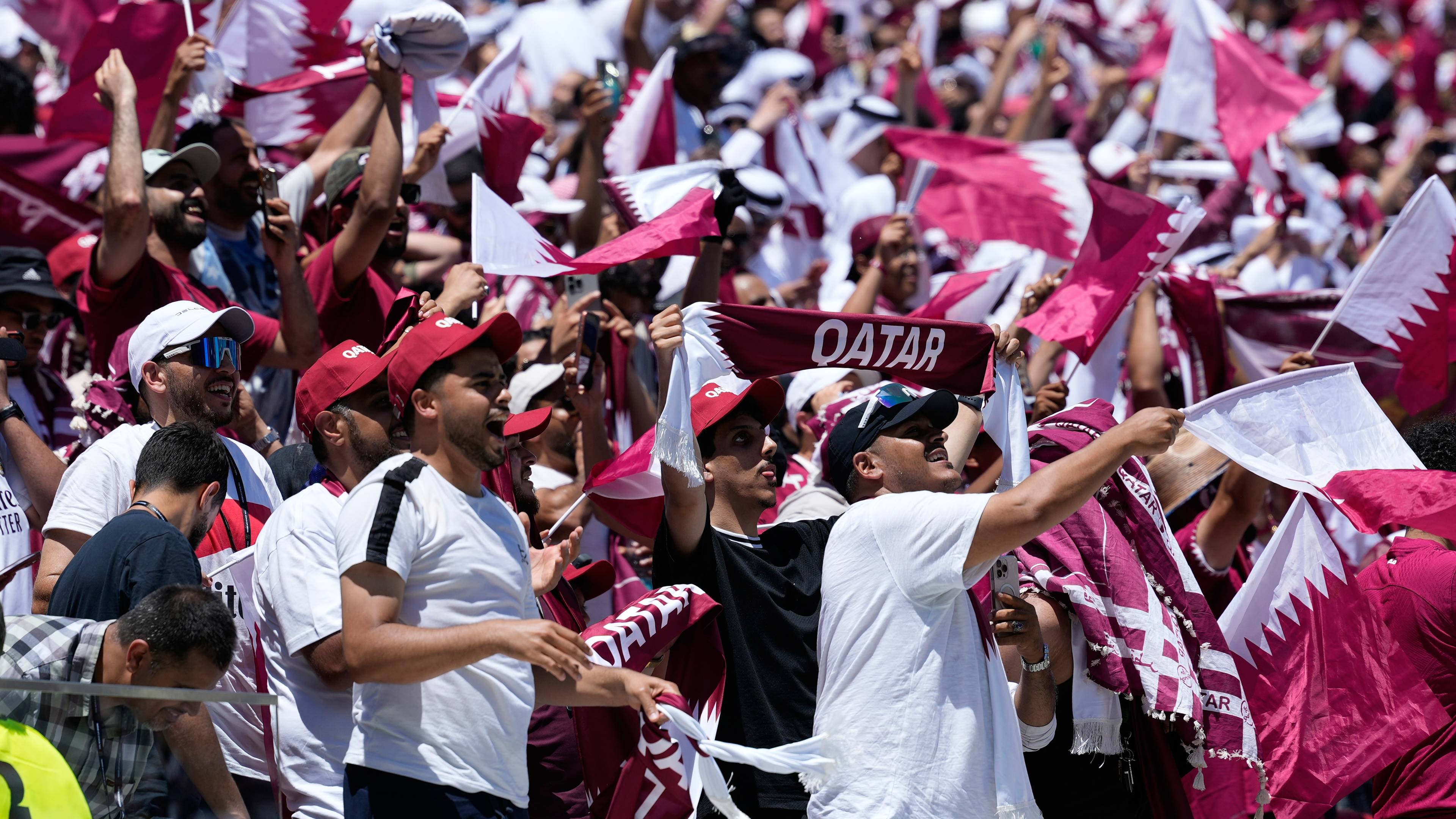 En stor gruppe tilskuere vifter med qatarske flagg. Flere av dem har på seg røde eller hvite caps og hvite t-skjorter. En person holder et banner med teksten "QATAR". Stemningen ser ut til å være festlig, med mange mennesker samlet. (Bildebeskrivelsen er laget av en KI-tjeneste)