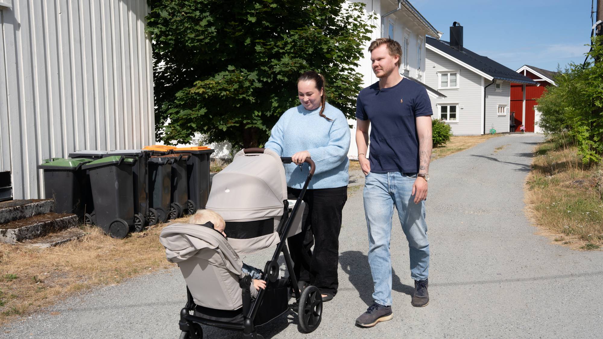 Kvinne og mann går tur på en grusvei med en barnevogn i et boligområde. (Bildebeskrivelsen er laget av en KI-tjeneste)