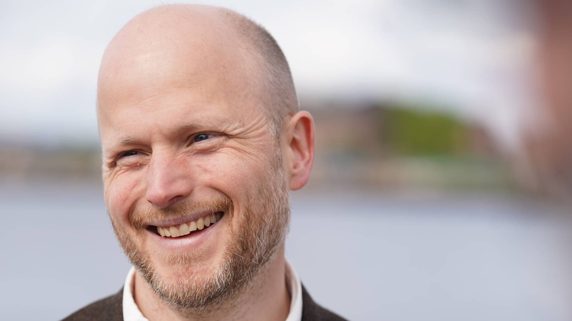 En mann med skallet hode og skjegg smiler mens han ser til siden. Han har på seg en mørk blazer over en hvit skjorte. I bakgrunnen er det uskarpt vann og grønne områder. (Bildebeskrivelsen er laget av en KI-tjeneste)