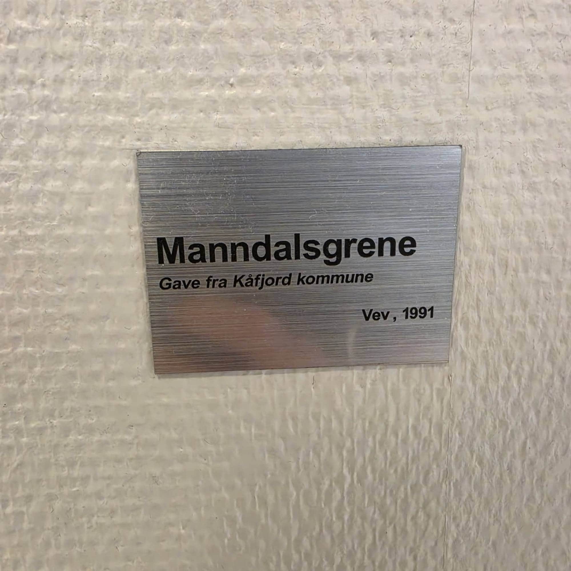 Metallskilt på vegg med teksten Manndalsgrene, gave fra Kåfjord kommune, Vev, 1991. (Bildebeskrivelsen er laget av en KI-tjeneste)