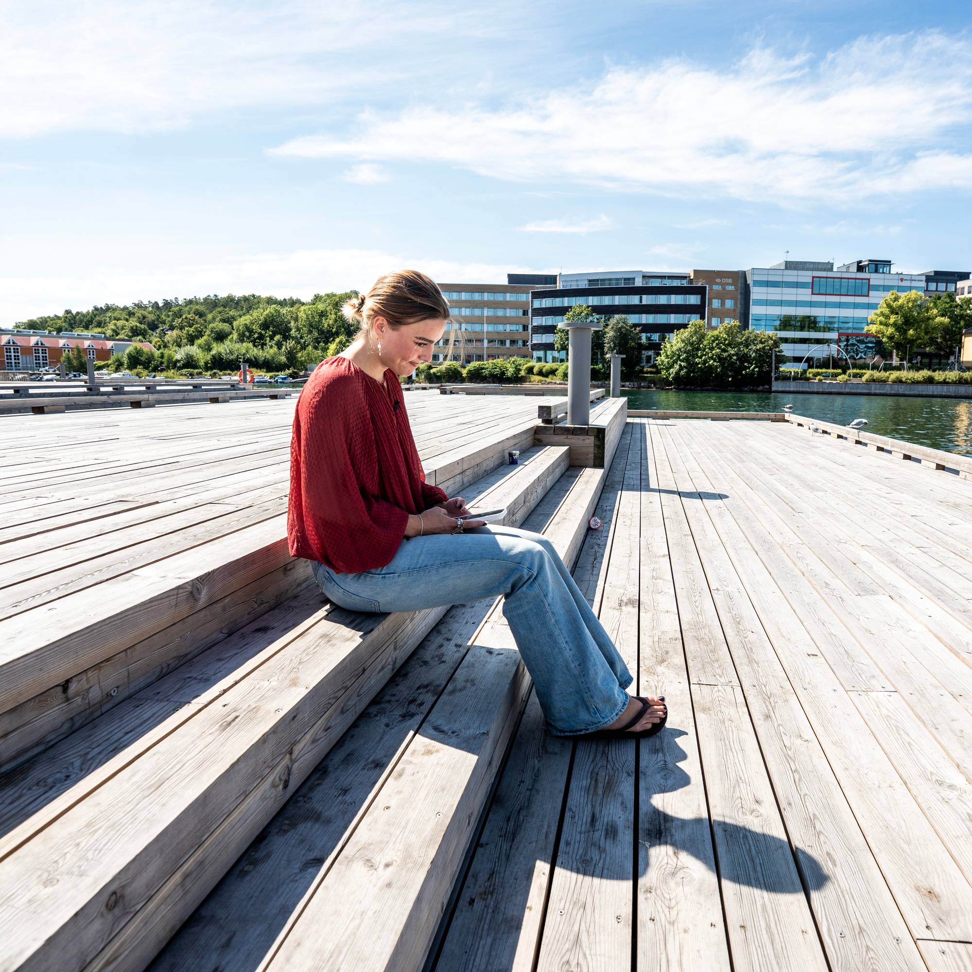 Kvinne sitter på en trebrygge ved vannet og ser ned på en mobiltelefon i hendene. 