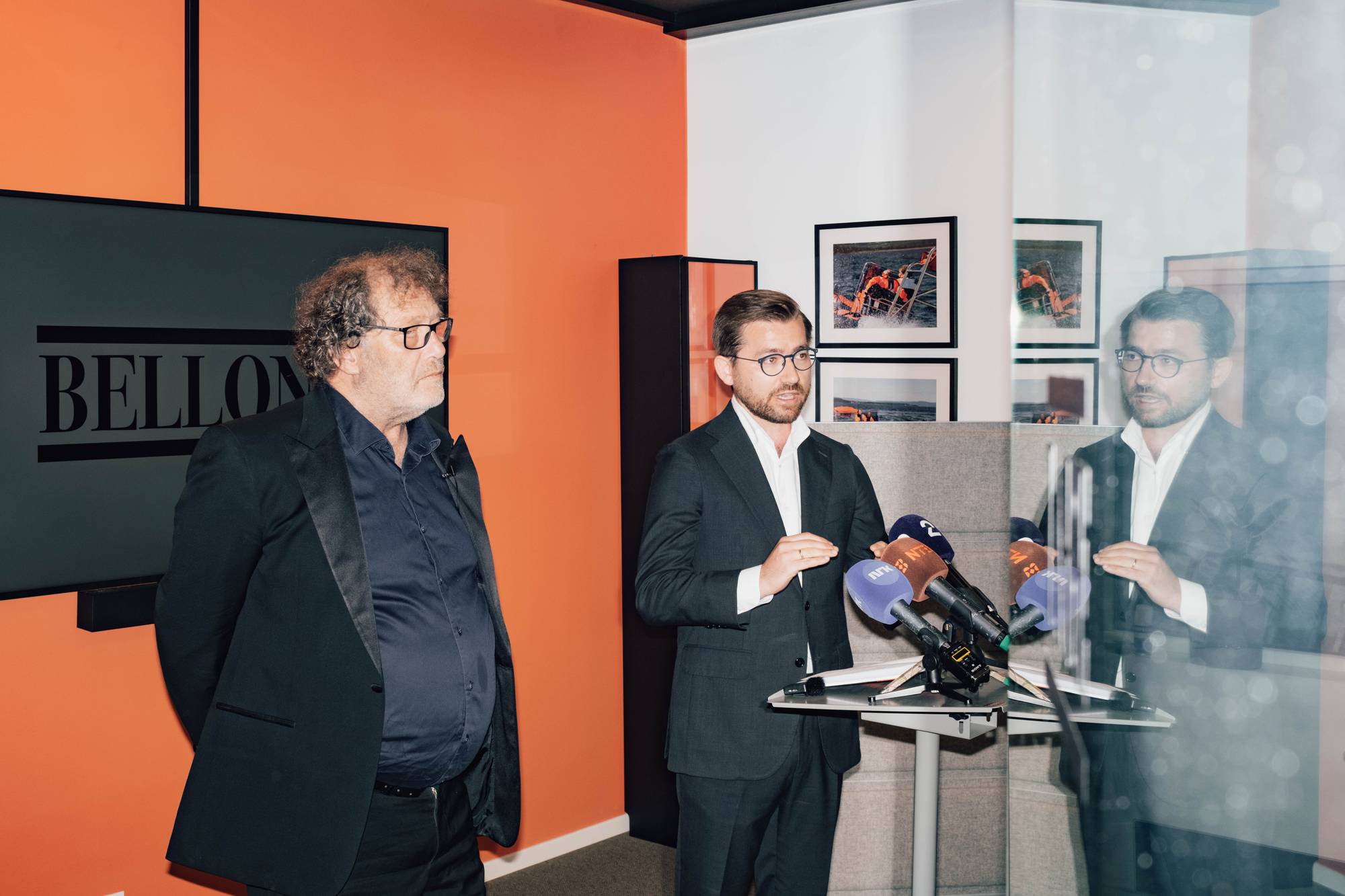 En mann i mørk dress snakker til et mikrofonstativ. En annen mann med briller står til venstre for ham. Bak dem er en stor skjerm med teksten "BELLONA". Rommet har oransje vegger og to bilder på veggen. (Bildebeskrivelsen er laget av en KI-tjeneste)