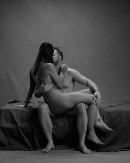 En kvinne med langt mørkt hår sitter på fanget til en mann med mørkt hår. Begge er nakne og kysser.