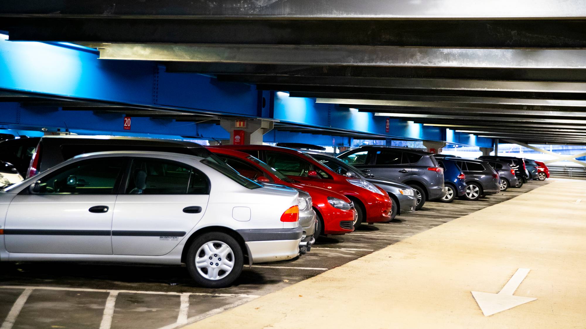 Biler parkert på rekke i et parkeringshus med blå bjelker i taket. (Bildebeskrivelsen er laget av en KI-tjeneste)