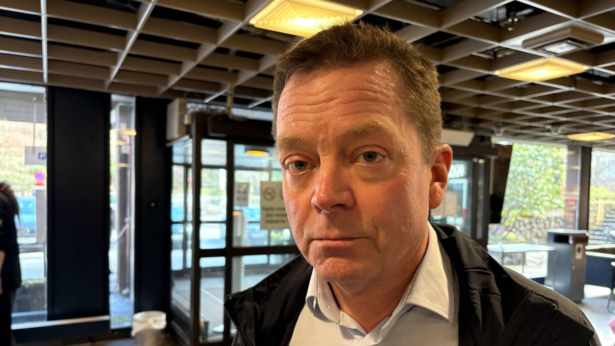 En mann står i en innendørs miljø, delvis vendt mot kameraet. Han har på seg en mørk jakke over en lys skjorte. I bakgrunnen sees andre personer og store vinduer som slipper inn naturlig lys. Det er et moderne rom med åpen dør til det utendørs området. (Bildebeskrivelsen er laget av en KI-tjeneste)