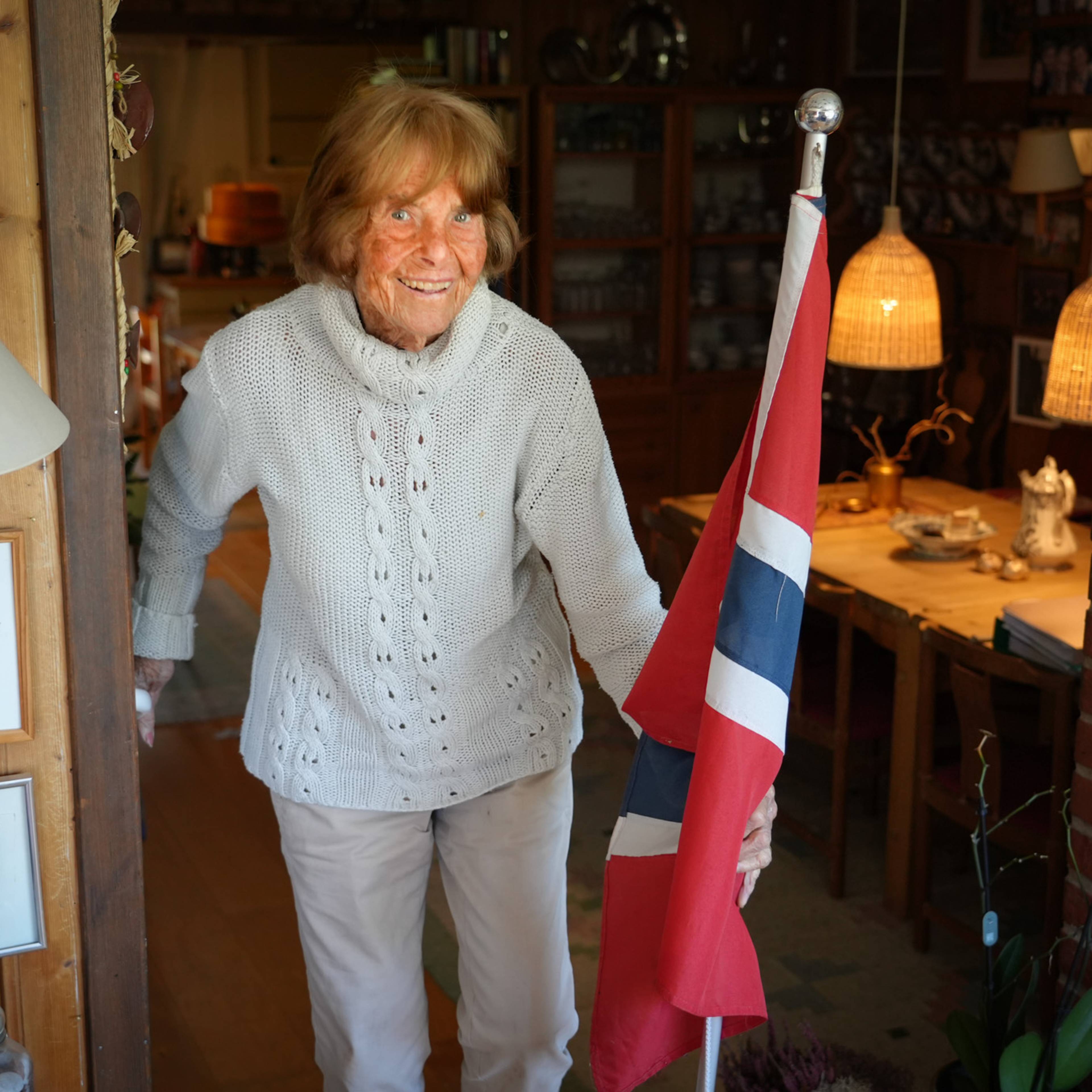 En eldre kvinne holder et norsk flagg. Hun er kledd i en lys strikkejakke og lyse bukser. Kvinnen står i en døråpning med et værelse bak seg. Både interiør og kunst på veggene er synlige. (Bildebeskrivelsen er laget av en KI-tjeneste)