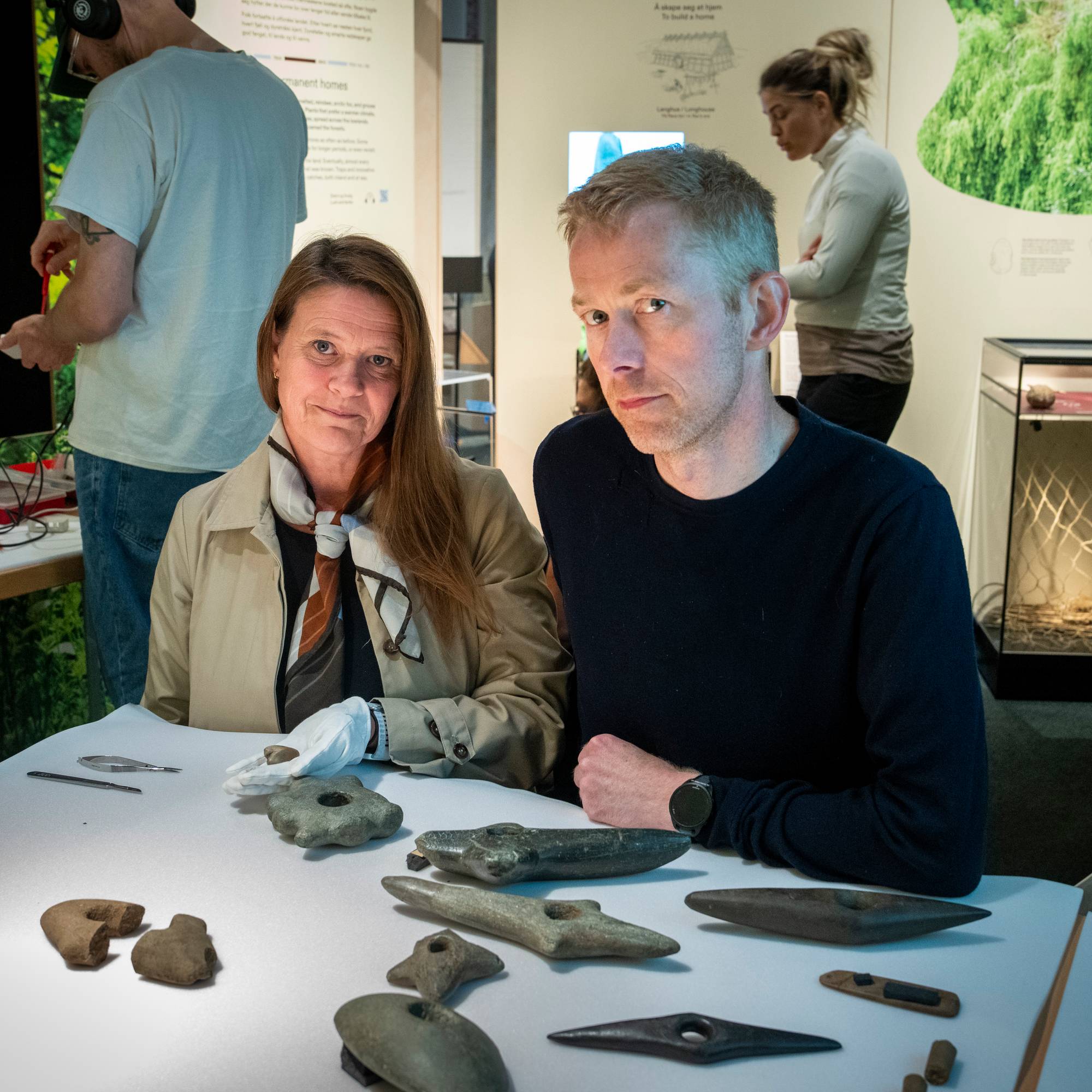 To personer sitter ved et bord med historiske gjenstander i et utstillingslokale. (Bildebeskrivelsen er laget av en KI-tjeneste)