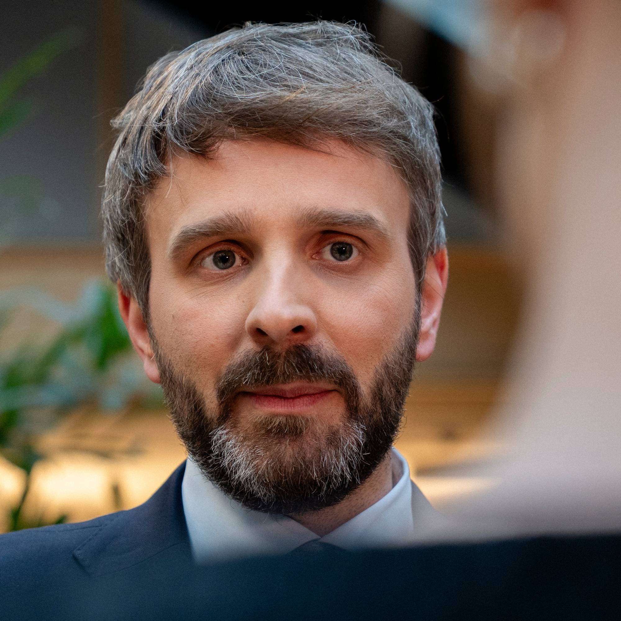 Et nærbilde viser en mann med grått skjegg og grått hår som ser mot kameraet. Mannen er iført en mørk dressjakke og hvit skjorte. Bak mannen er en utydelig person og planter. (Bildebeskrivelsen er laget av en KI-tjeneste)
