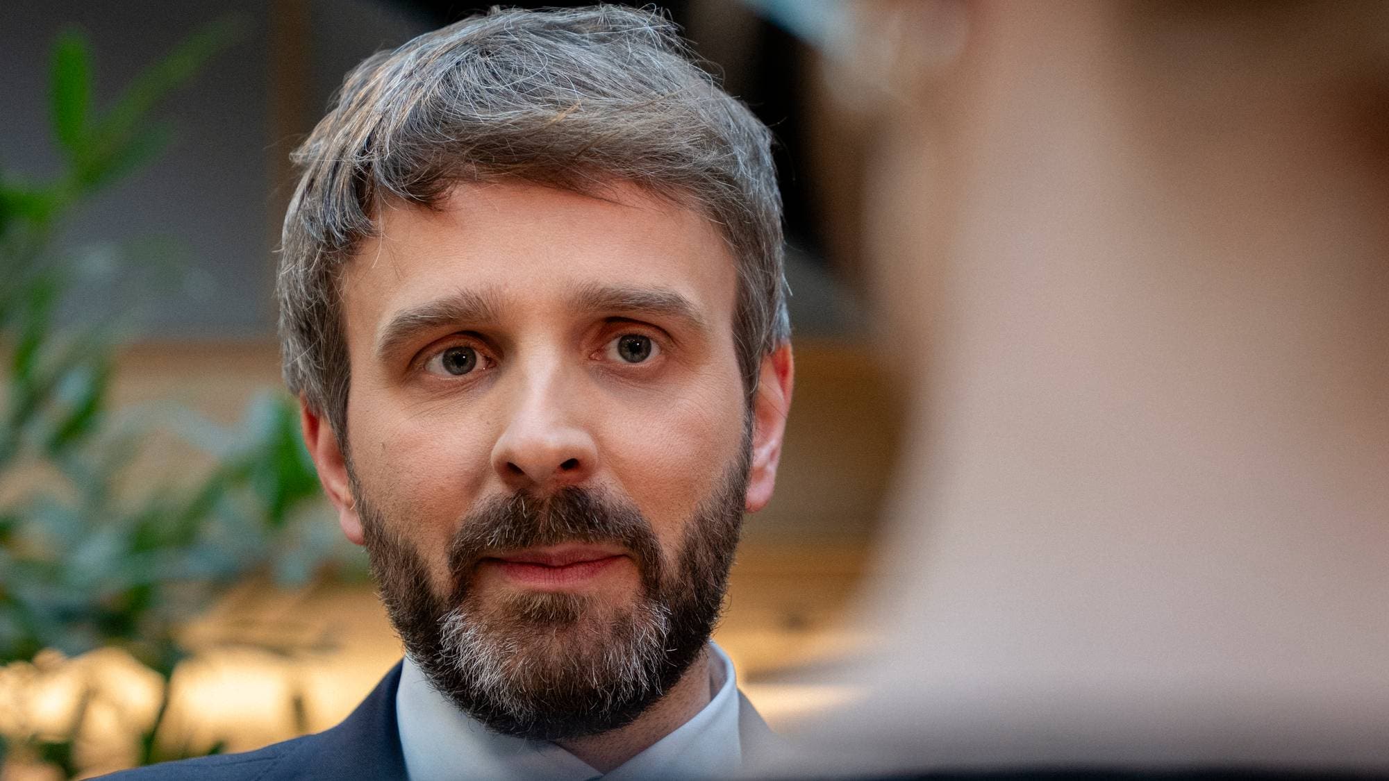 Et nærbilde viser en mann med grått skjegg og grått hår som ser mot kameraet. Mannen er iført en mørk dressjakke og hvit skjorte. Bak mannen er en utydelig person og planter. (Bildebeskrivelsen er laget av en KI-tjeneste)