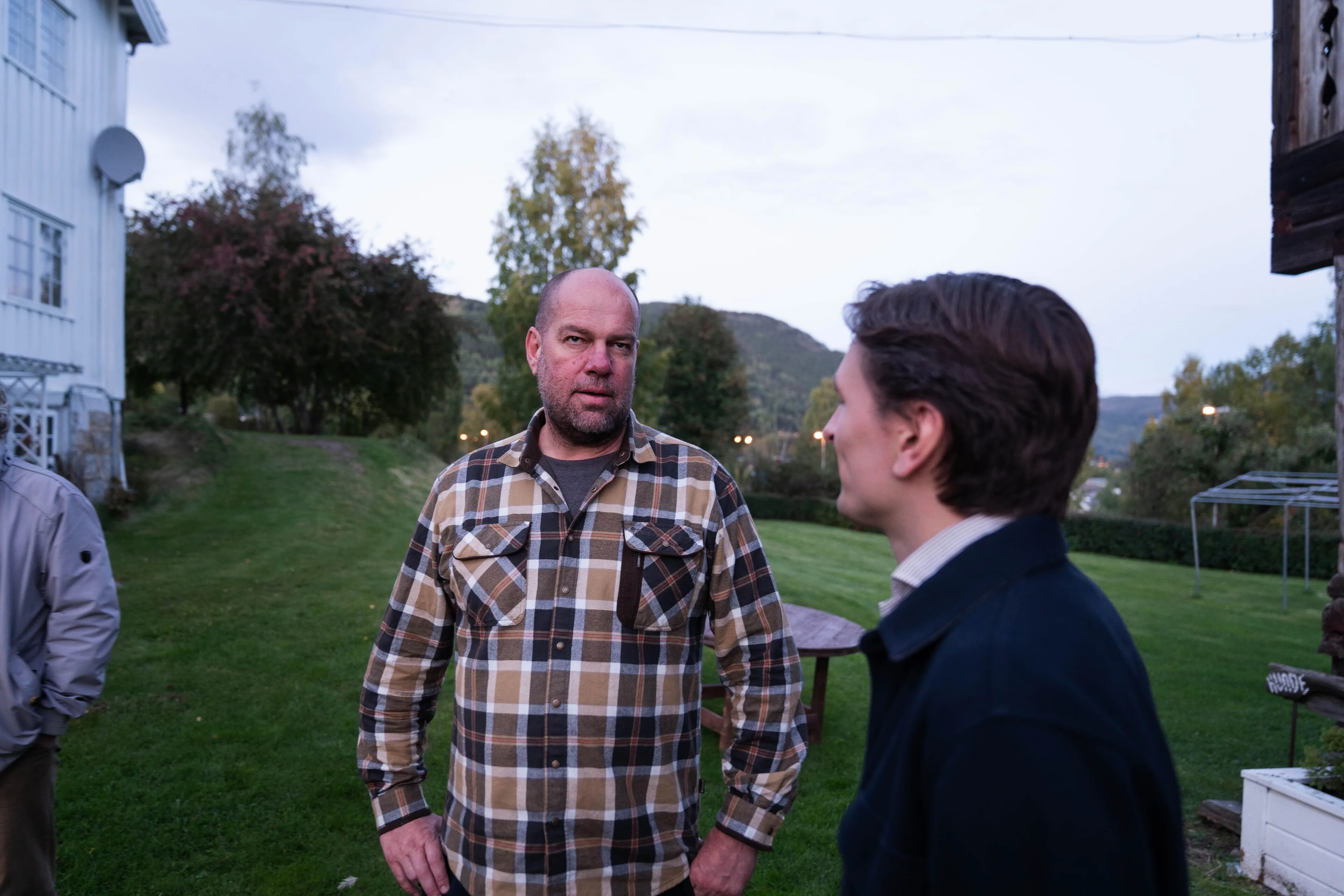 To menn står utendørs og snakker sammen på en gressplen foran et trehus. (Bildebeskrivelsen er laget av en KI-tjeneste)
