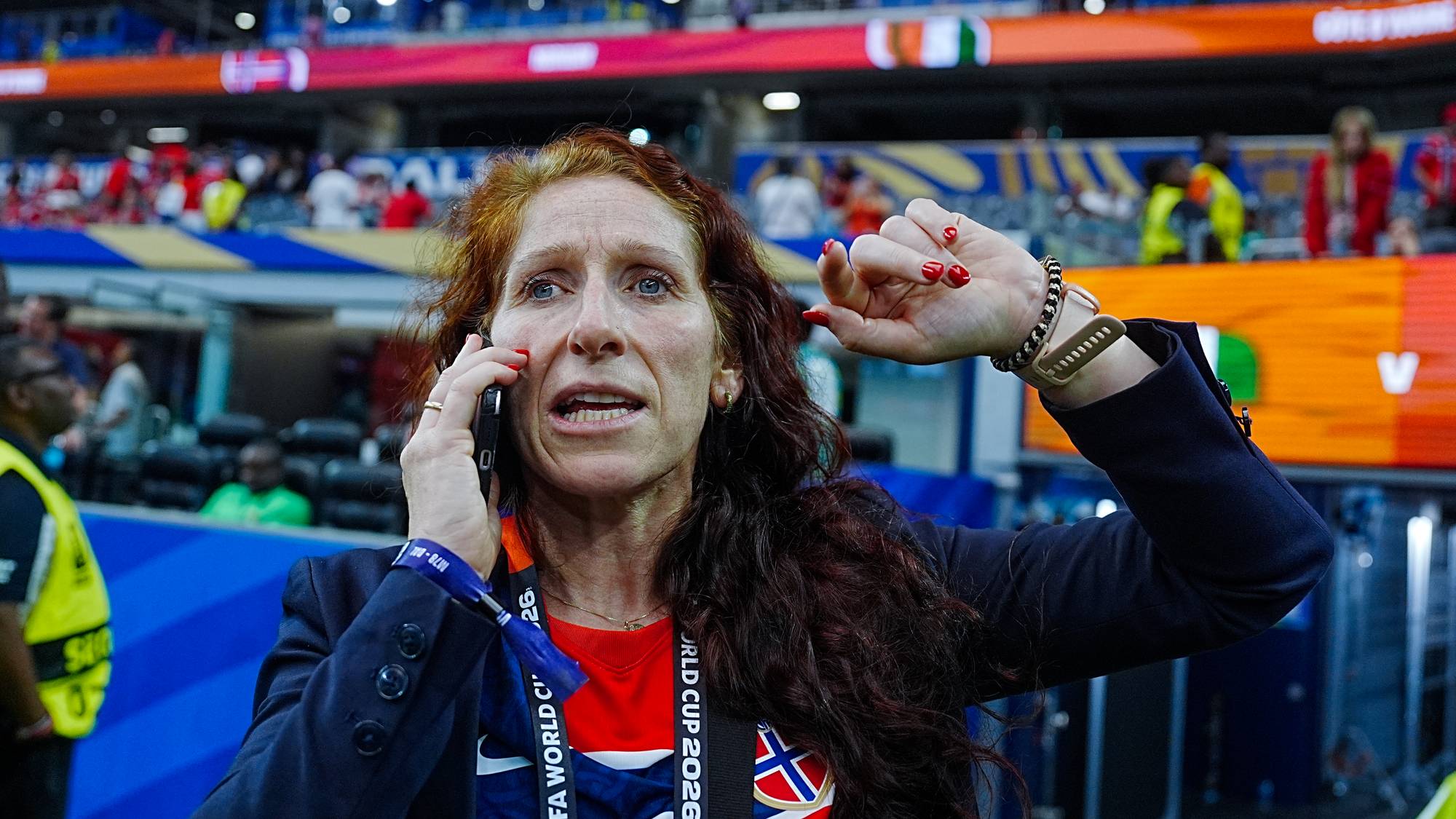 Kvinne med norsk landslagsdrakt snakker i mobiltelefon på en fotballstadion. (Bildebeskrivelsen er laget av en KI-tjeneste)