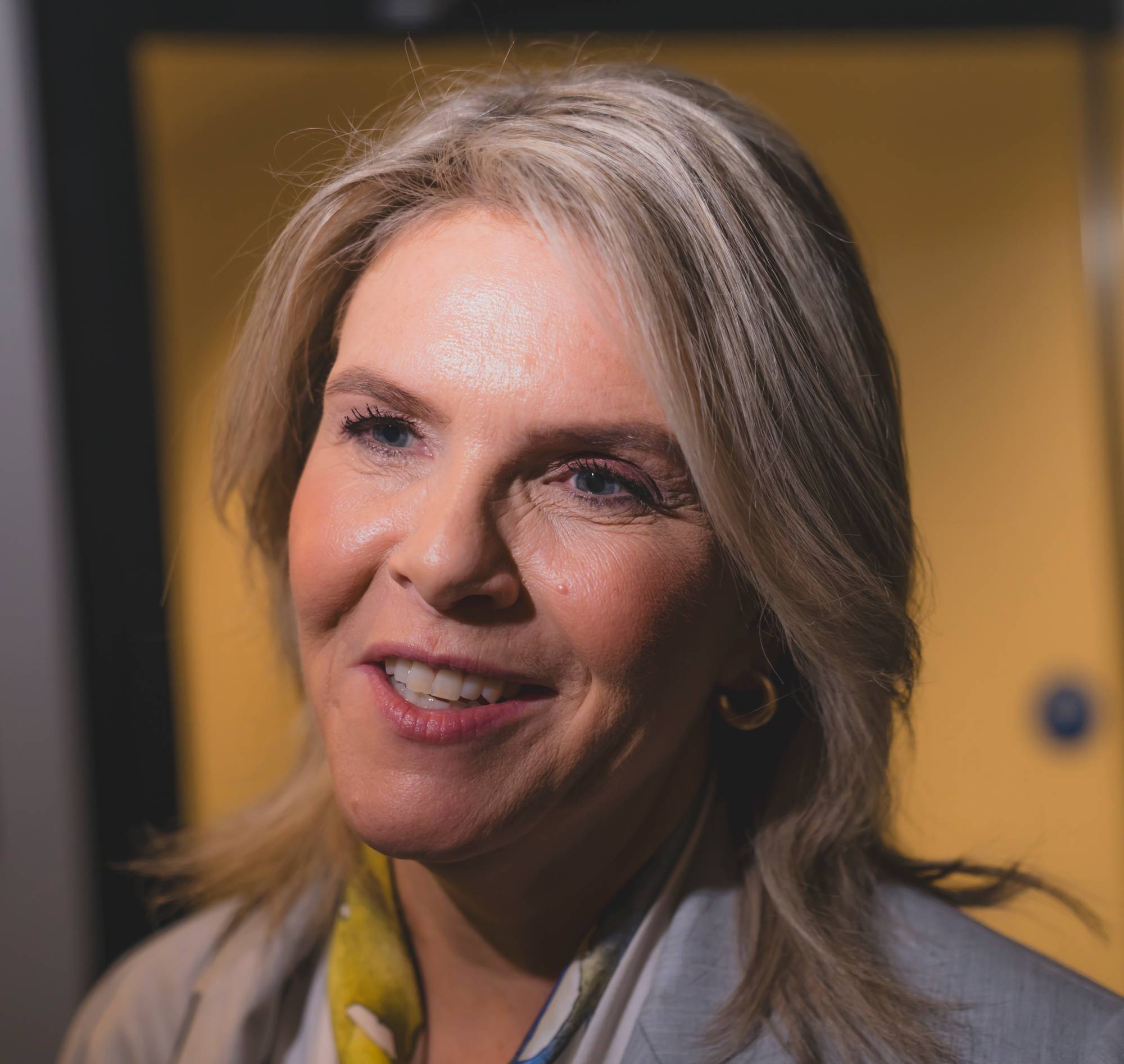 Portrett av Sylvi Listhaug, som smiler mot kamera. Bakgrunnen er en gul vegg. (Bildebeskrivelsen er laget av en KI-tjeneste)