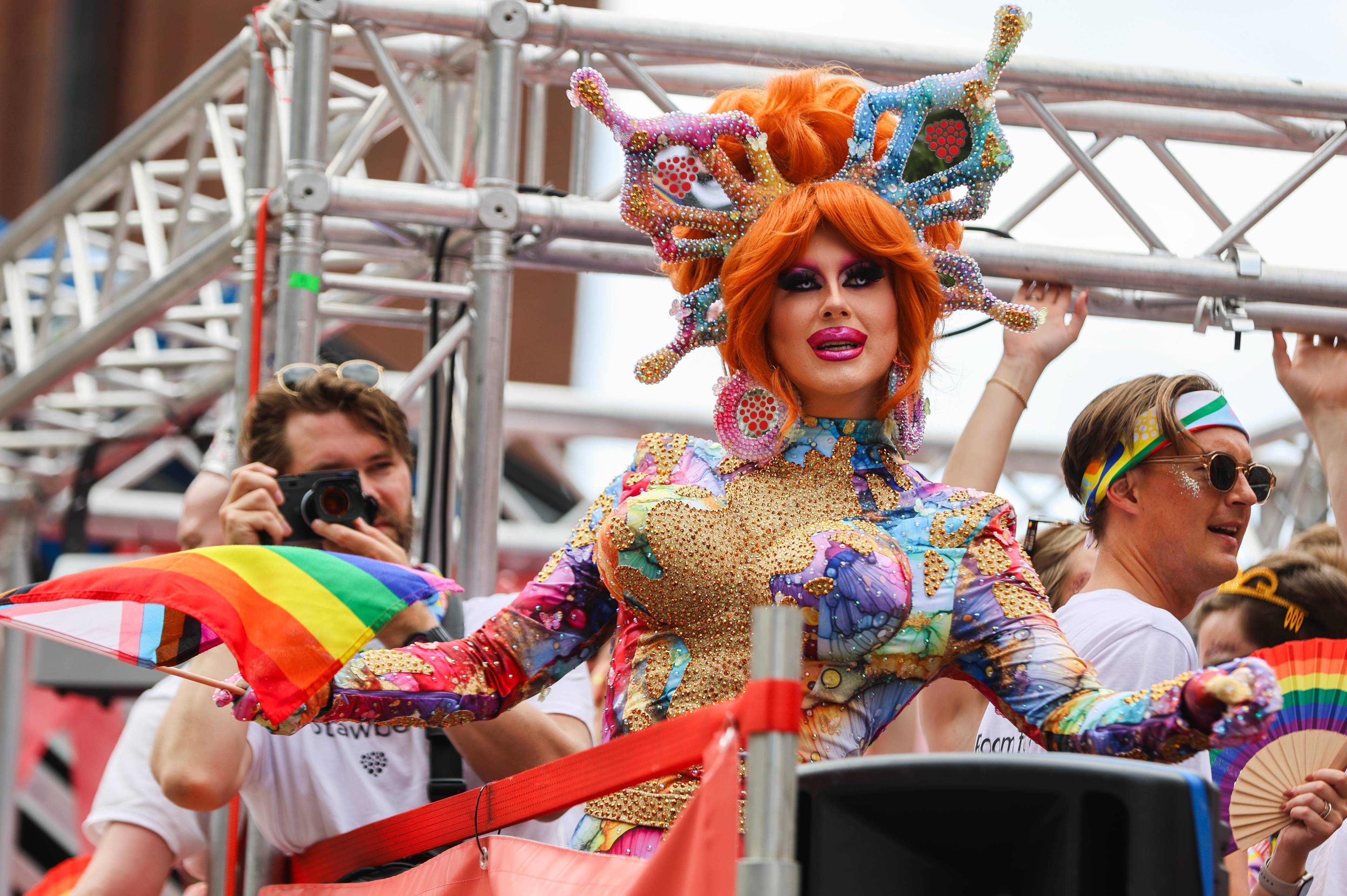 En dragartist i fargerikt kostyme og hodeplagg deltar i en Pride-parade på en bil. (Bildebeskrivelsen er laget av en KI-tjeneste)