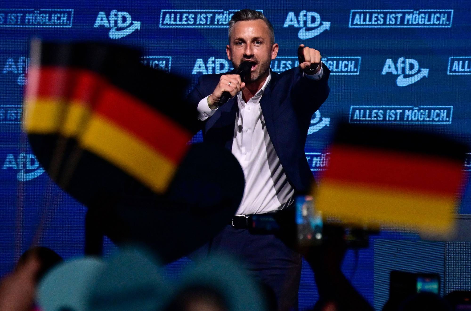 mann snakker i mikrofon på scene med flagg i forgrunn