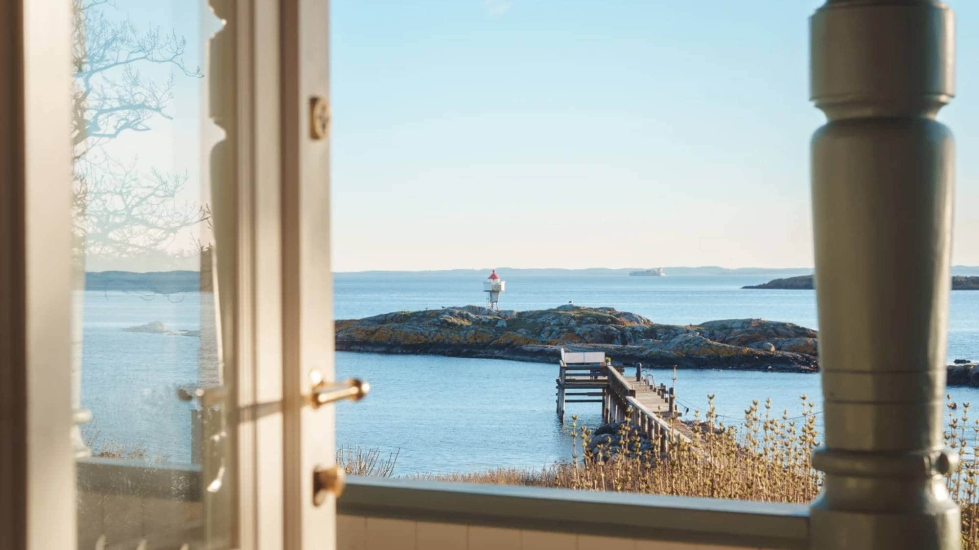 Et åpent hvitt vindu viser en skjærgård med en gangbro og et fyrtårn på en holme. I bakgrunnen er en stor lekter på havet under en klar blå himmel. Lyset faller på den vakre kystlinjen. (Bildebeskrivelsen er laget av en KI-tjeneste)