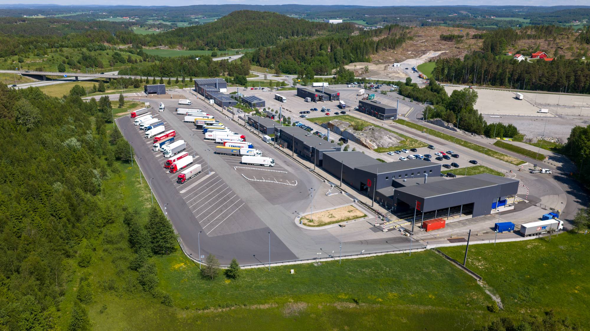 Et stort antall lastebiler er parkert på en rasteplass med nærliggende bygninger. I bakgrunnen sees en motorvei og et skogkledd landskap. Omgivelsene er grønne med noen flekker med bar mark. (Bildebeskrivelsen er laget av en KI-tjeneste)