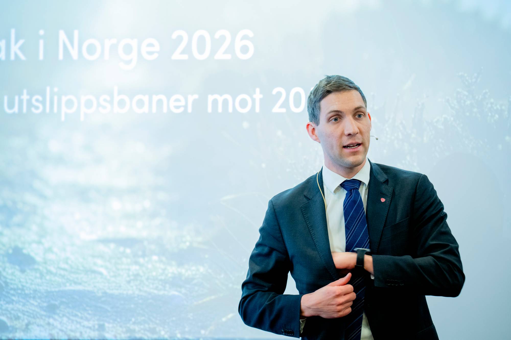 Ung mann med mørkt hår i dress foran en lyseblå bakgrunn med tekst
