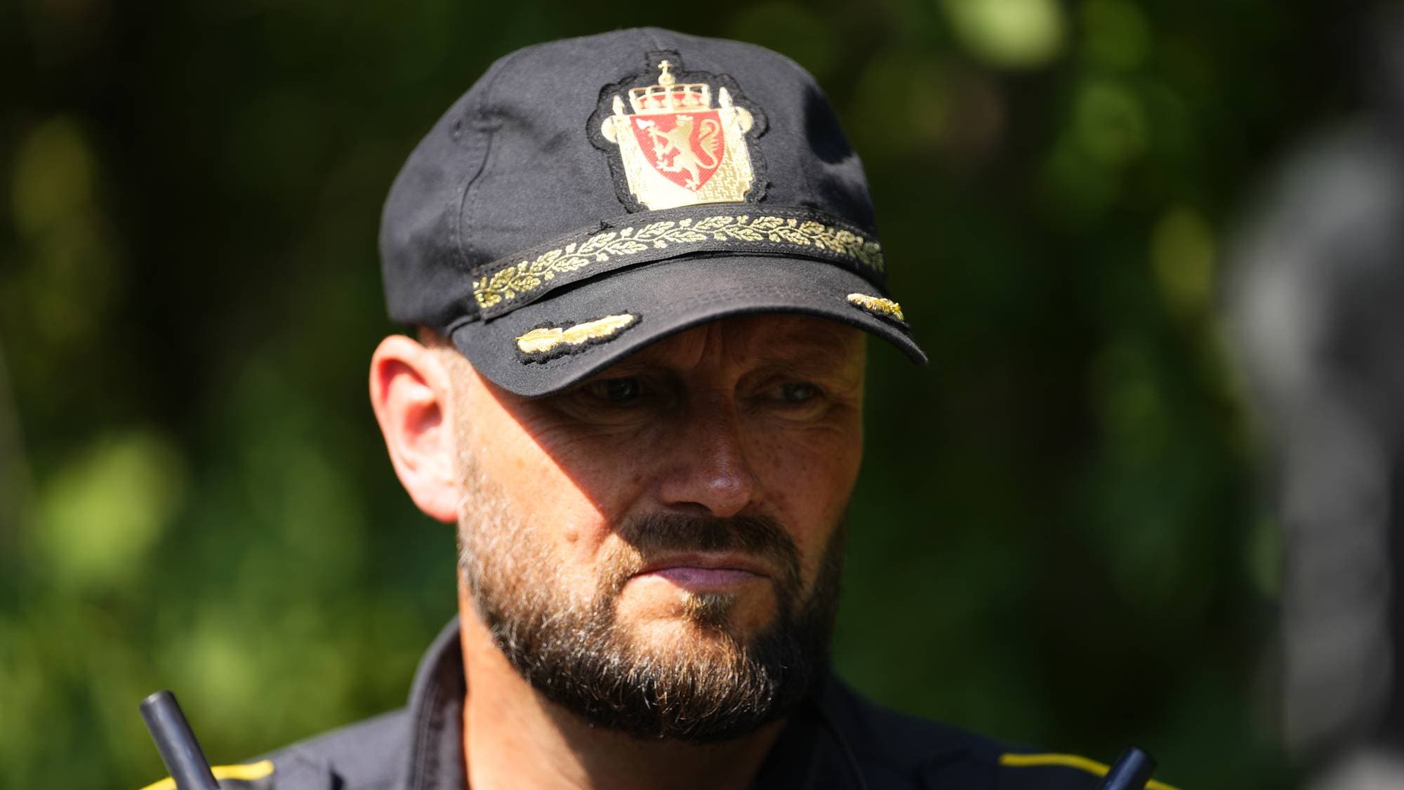 Politibetjent i uniform med caps med riksvåpenet. Utendørs bakgrunn med grønne blader. (Bildebeskrivelsen er laget av en KI-tjeneste)
