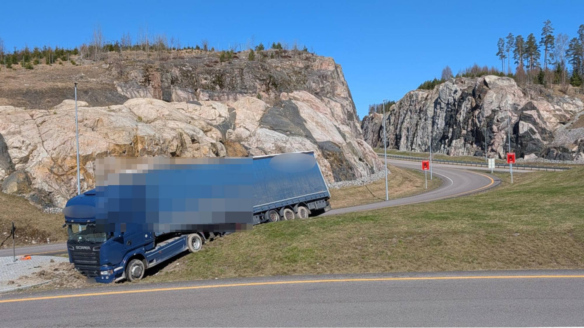 n blå semitrailer har kjørt av veien og står i en skråning ved en fjellside. Veien er asfaltert og svinger til høyre. Det er grønt gress på sidene av veien og fjellknauser og trær i bakgrunnen. Himmelen er blå og skyfri. (Bildebeskrivelsen er laget av en KI-tjeneste)
