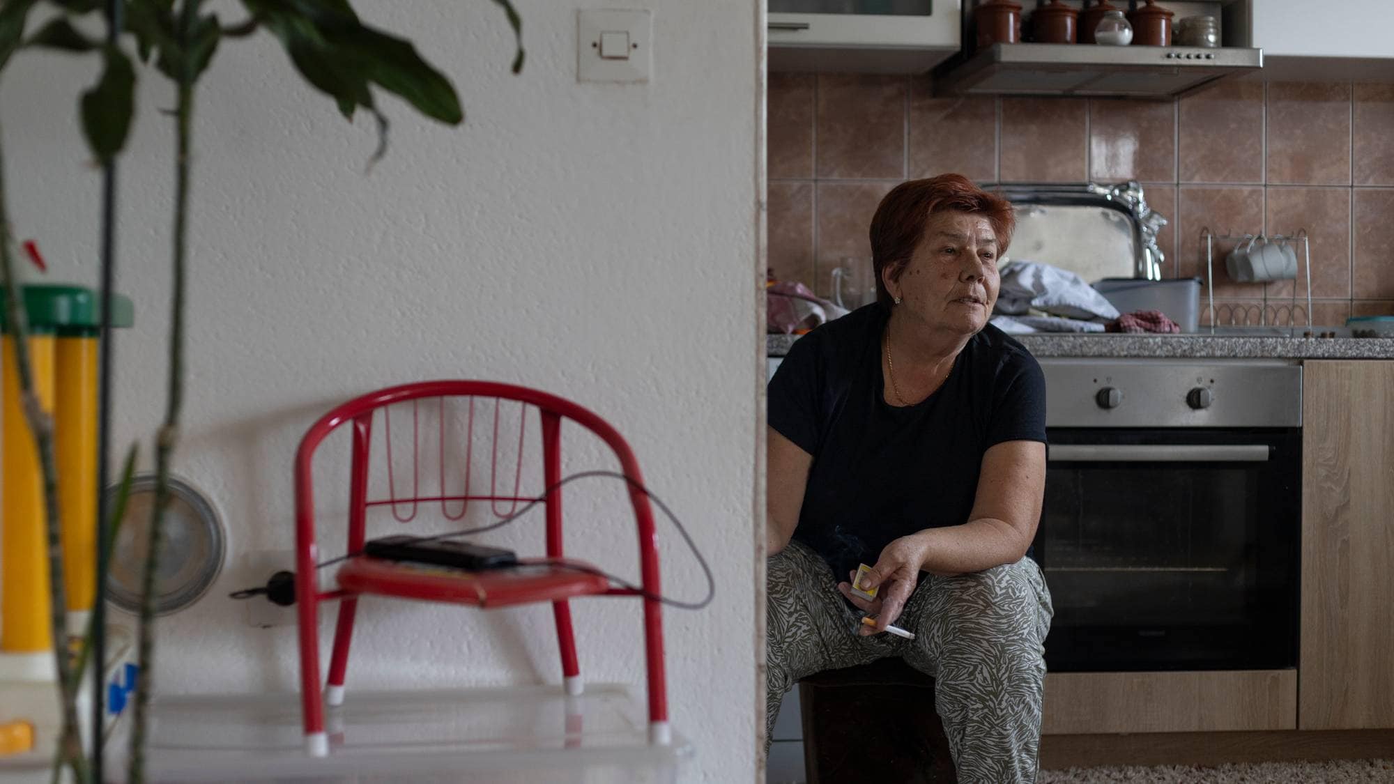 En eldre kvinne sitter på en krakk på kjøkkenet. Hun har en sigarett i hånden og kikker ut mot siden