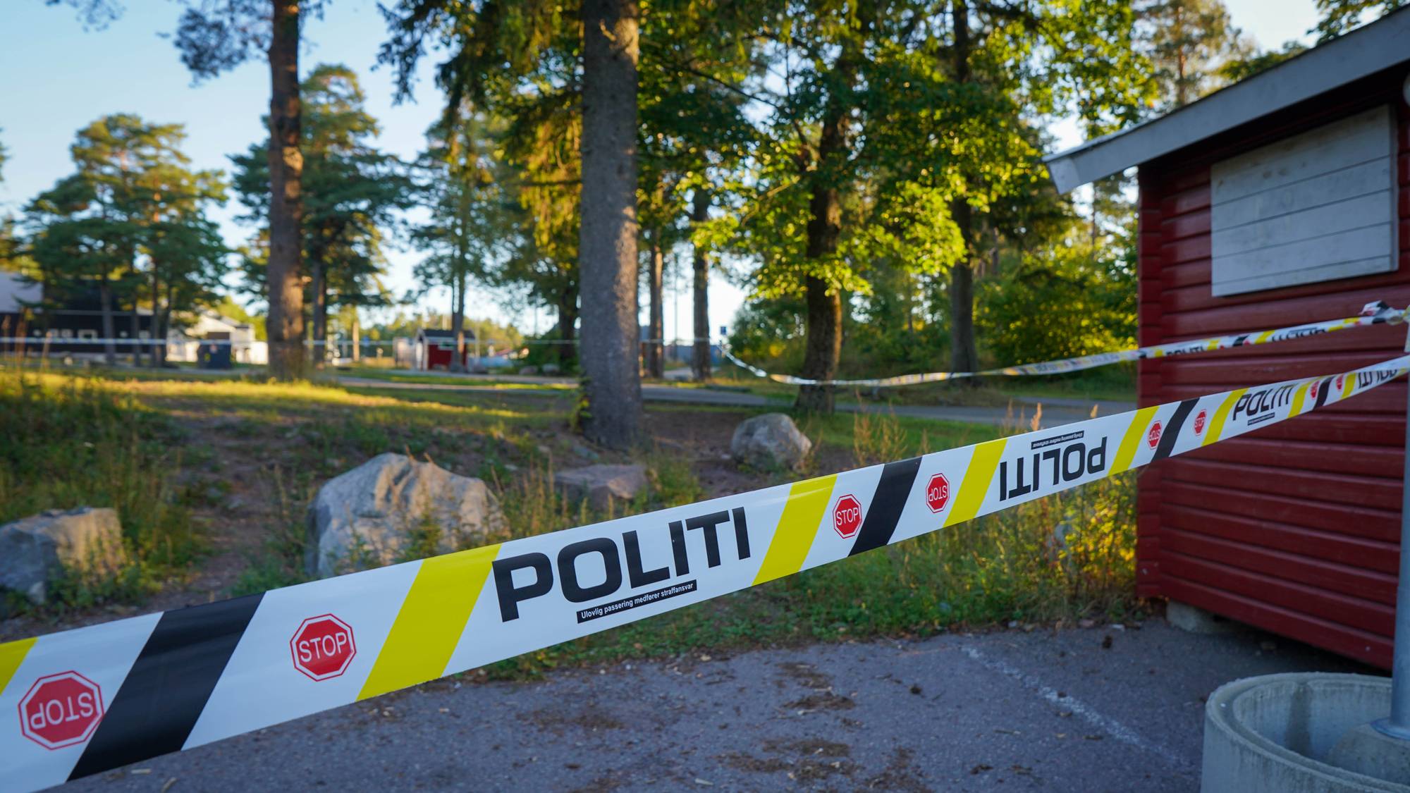Politi-sperrebånd som sperrer av deler av et naturområde. Et rødt bygg skimtes til høyre i bildet.