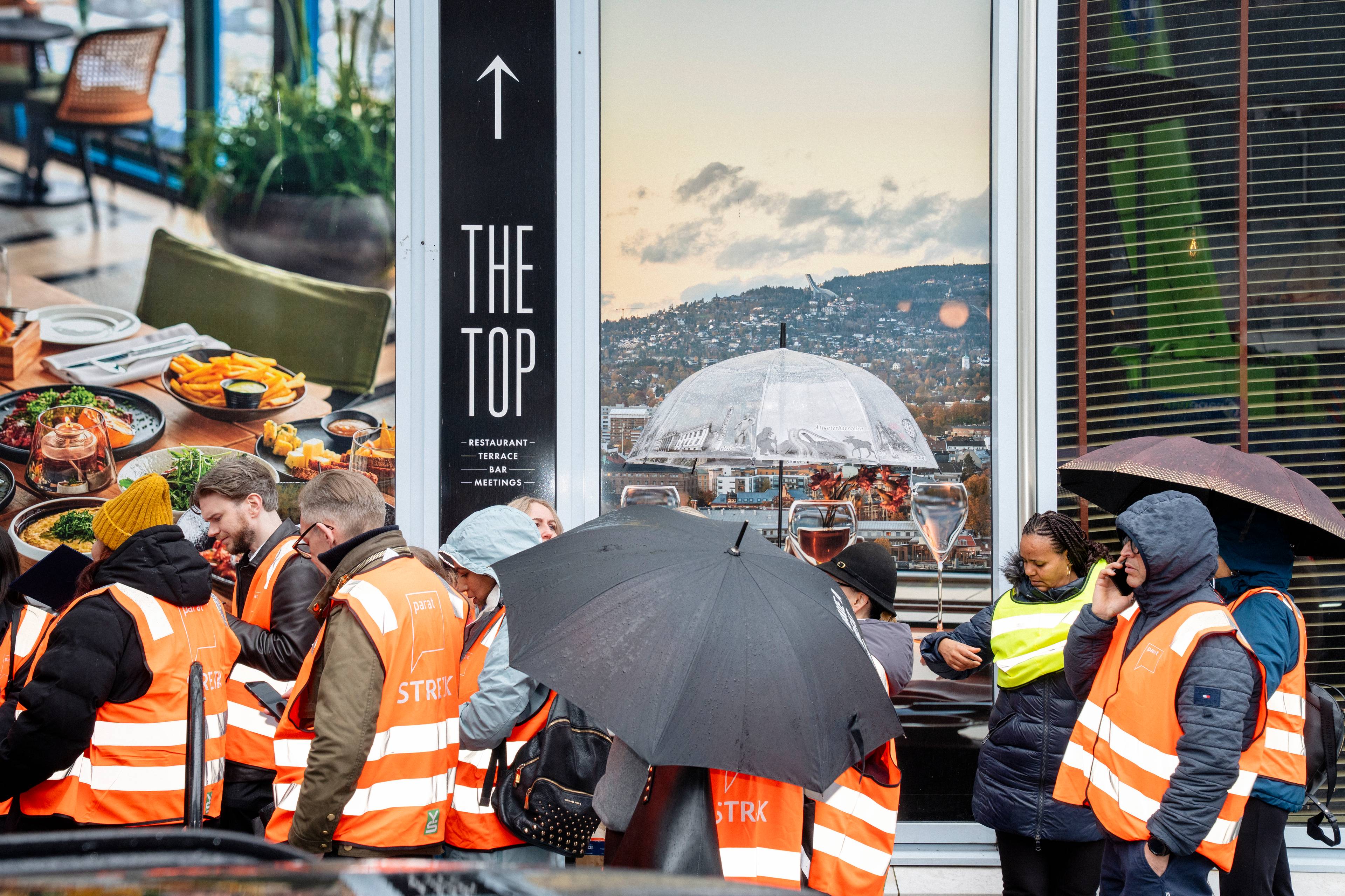En gruppe mennesker med oransje refleksvester står samlet ute i regnvær. De holder paraplyer for å beskytte seg mot nedbøren. En av personene snakker i telefonen. Bak dem er det store vinduer med bilder og tekst. (Bildebeskrivelsen er laget av en KI-tjeneste)
