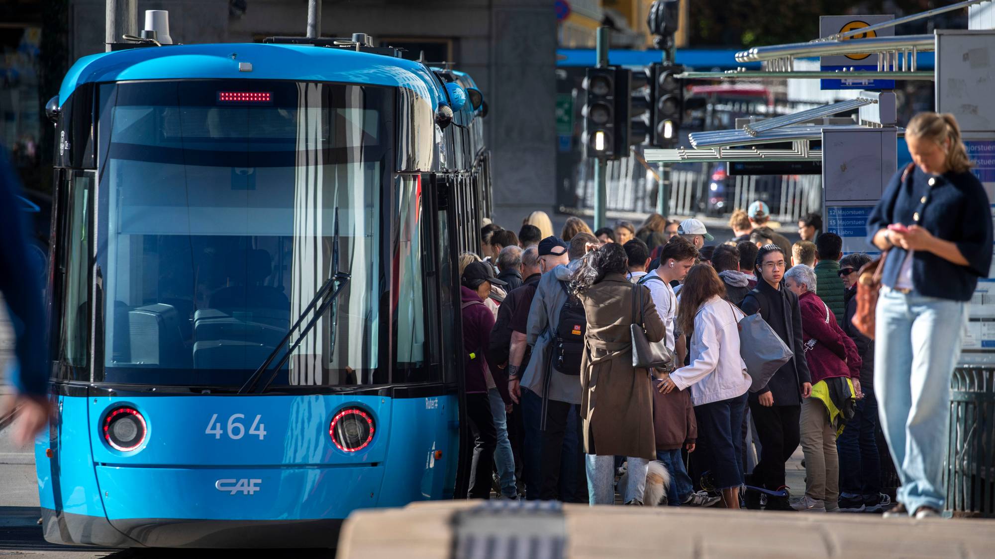 Folk står på perrongen og venter på en blå bytrikk i Oslo. (Bildebeskrivelsen er laget av en KI-tjeneste)