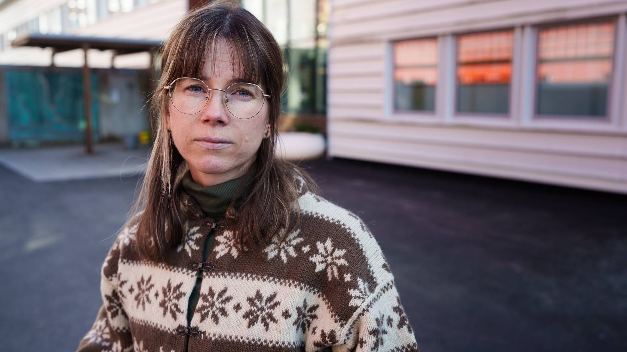 En person står i forgrunnen iført en strikkegenser med mønster, delvis skjult ansikt. Bakgrunnen viser en bygning med vinduer som reflekterer lyset fra omgivelsene. Det er en asfaltlagt overflate foran personen, som tyder på et utendørs miljø. Bildet har en klar og lys atmosfære. (Bildebeskrivelsen er laget av en KI-tjeneste)