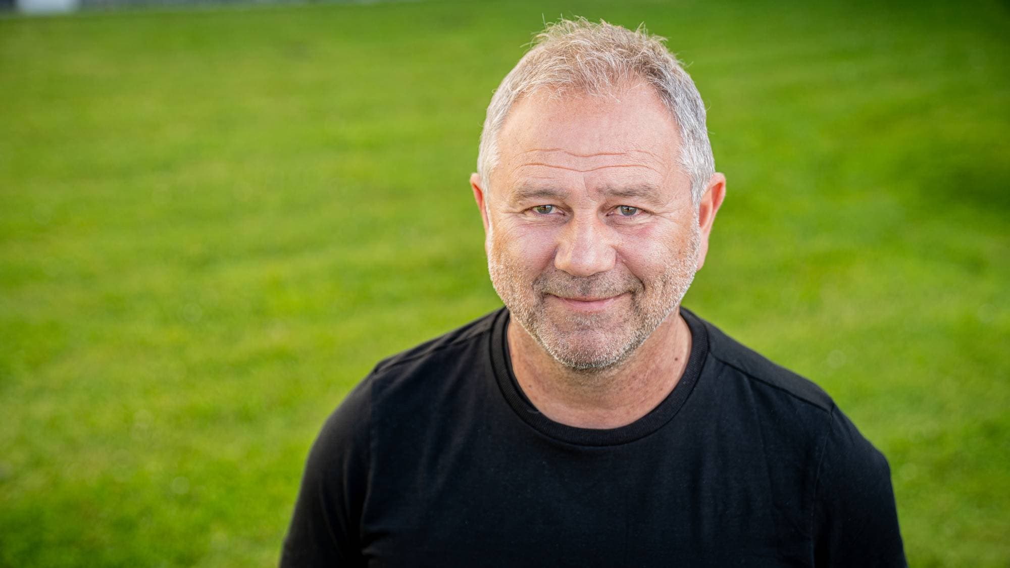 Mann med grålig hår og skjegg i svart t-skjorte fotografert utendørs foran en gressplen. (Bildebeskrivelsen er laget av en KI-tjeneste)