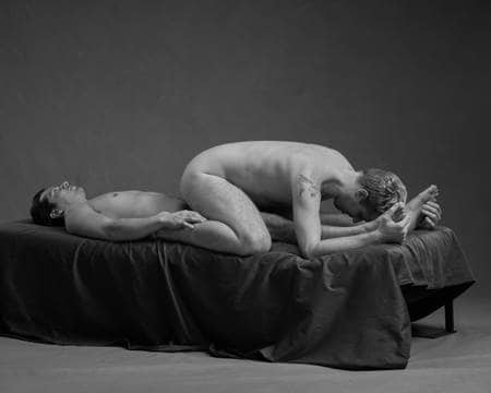 En naken mann ligger på ryggen i senga mens han holder en anen naken mann på leggen som sitter over ham med hodet ned mot føttene hans