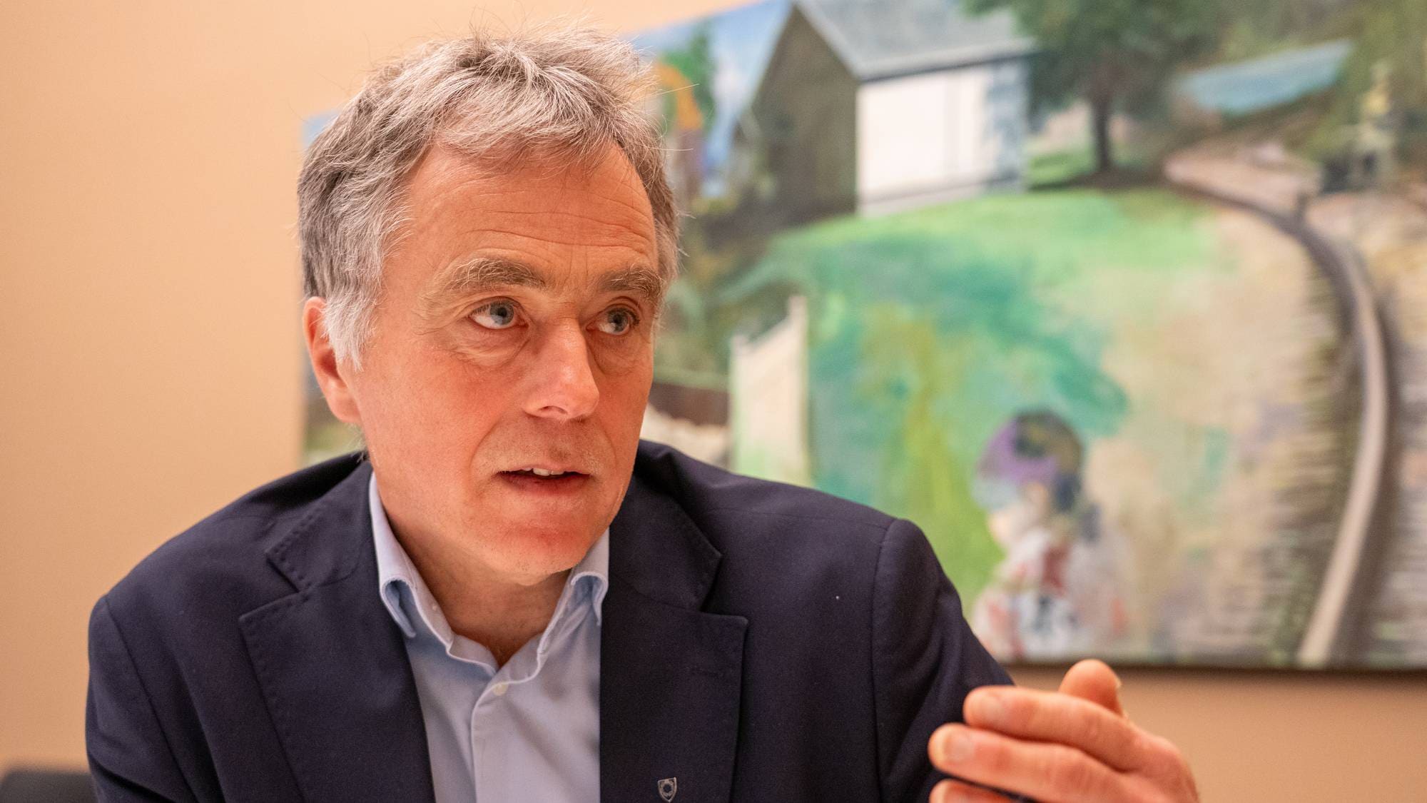 En mann i mørk dressjakke og lys skjorte sitter og snakker. Bak ham henger et fargerikt maleri av et landskap med et hus og en person. Mannens hånd er hevet som om han gestikulerer under samtalen. Bakgrunnen er en nøytral vegg. (Bildebeskrivelsen er laget av en KI-tjeneste)