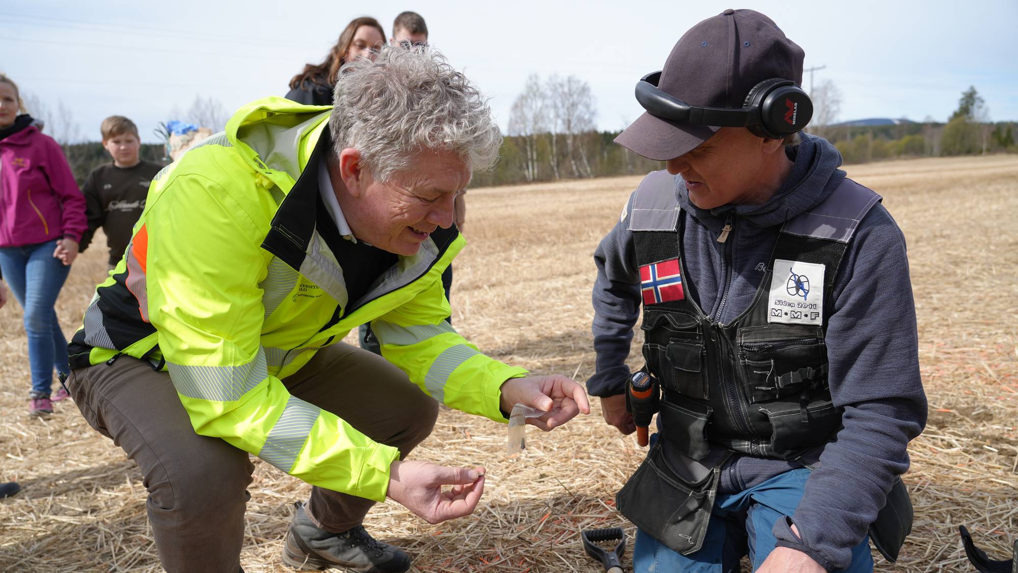 To personer på et jorde, en viser frem et lite funn. Den ene personen har på seg en gul refleksvest og hvit skjorte. Den andre personen har på seg en vest med norske flagg og et merke. En spade ligger i gresset. Bak dem står flere andre personer.