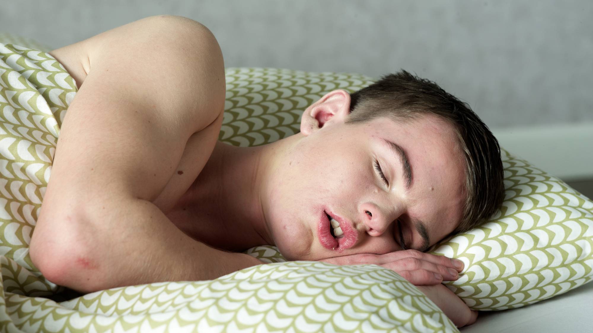 En ung mann sover med hodet på en pute med grønt mønster. Han er tildekket av en dyne. (Bildebeskrivelsen er laget av en KI-tjeneste)
