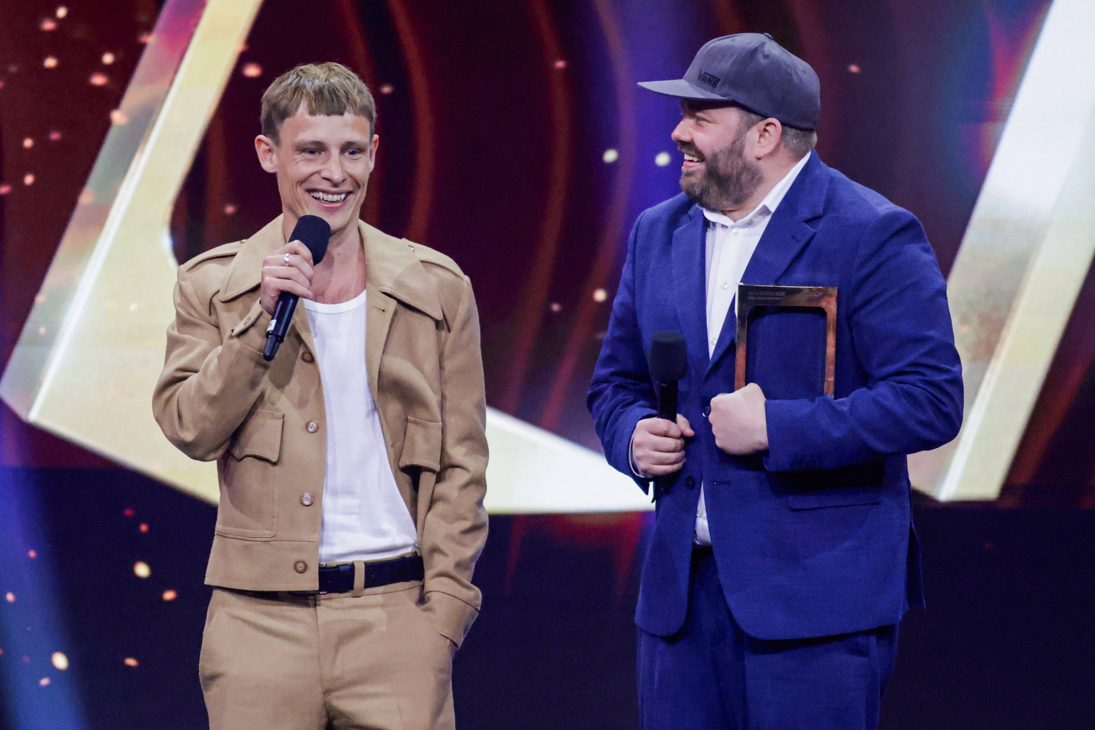 To menn står på en scene med en pris. Den ene mannen holder en mikrofon og smiler. Den andre mannen har på seg en blå dress og cap, og holder en pris i hånden. (Bildebeskrivelsen er laget av en KI-tjeneste)