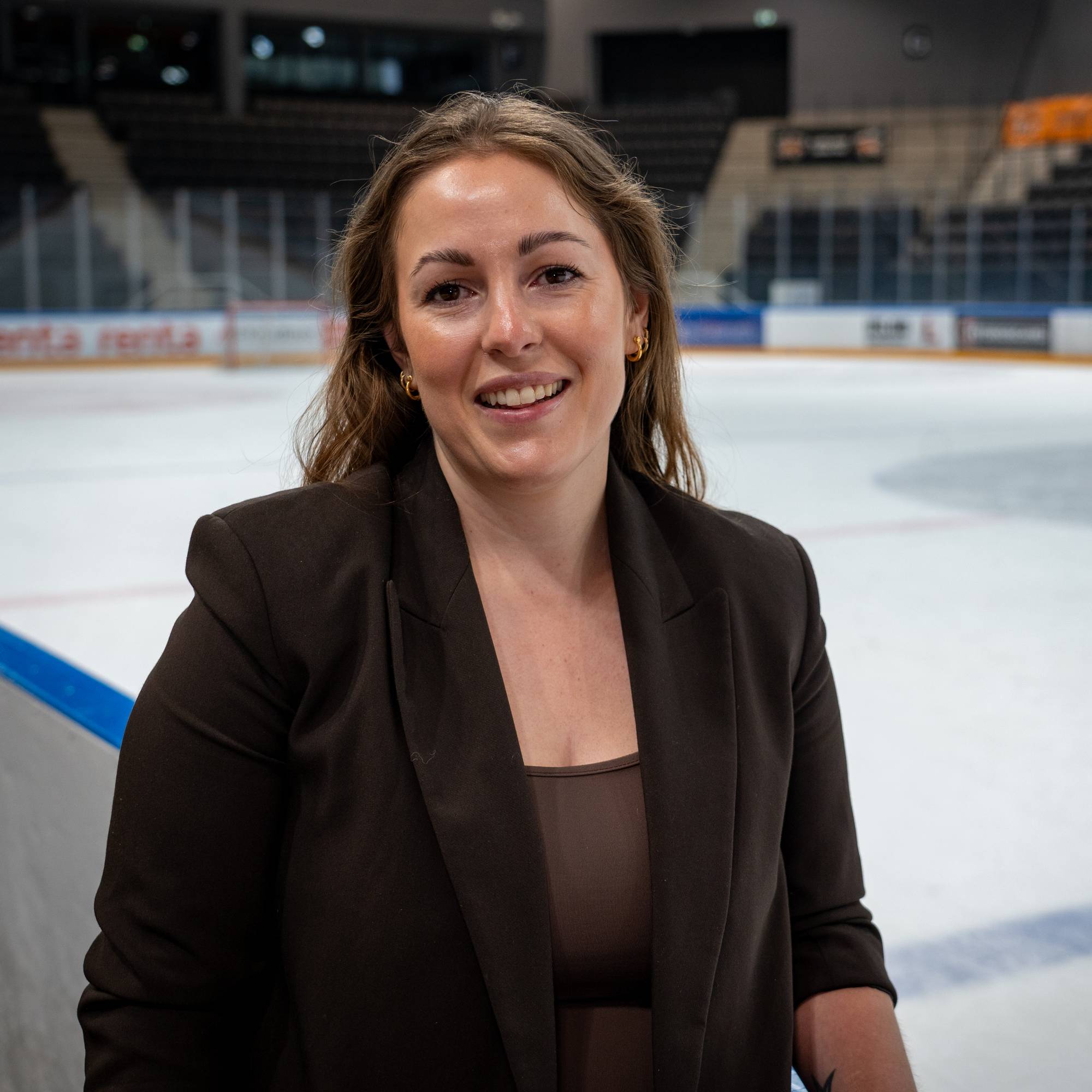 Kvinne i mørk blazer står foran en ishockeybane i en tom ishall. (Bildebeskrivelsen er laget av en KI-tjeneste)