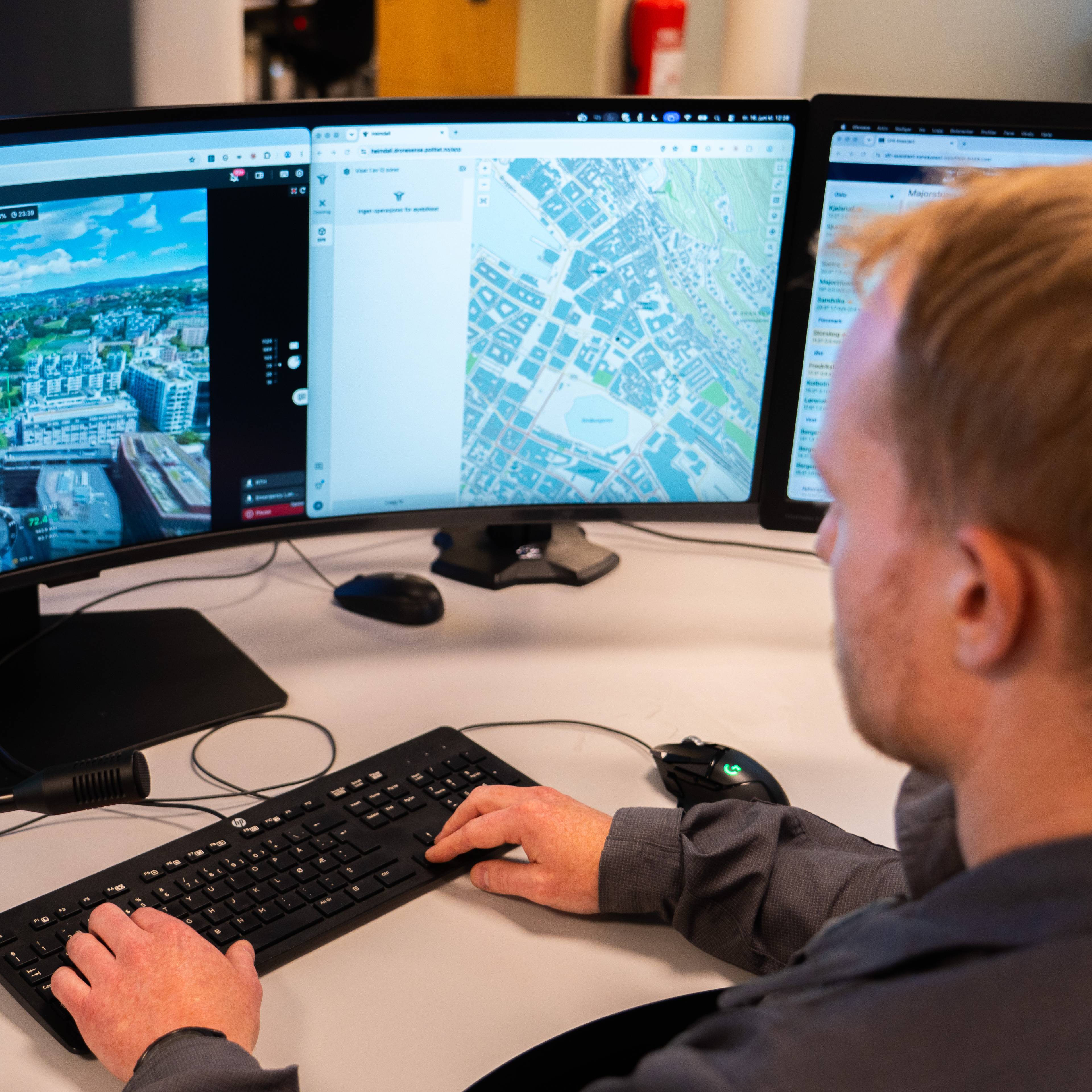 Mann ser på en buet skjerm som viser dronebilder av bylandskap og kart. Han sitter ved et skrivebord med tastatur og mus. En mikrofon er festet til et stativ på skrivebordet. (Bildebeskrivelsen er laget av en KI-tjeneste)
