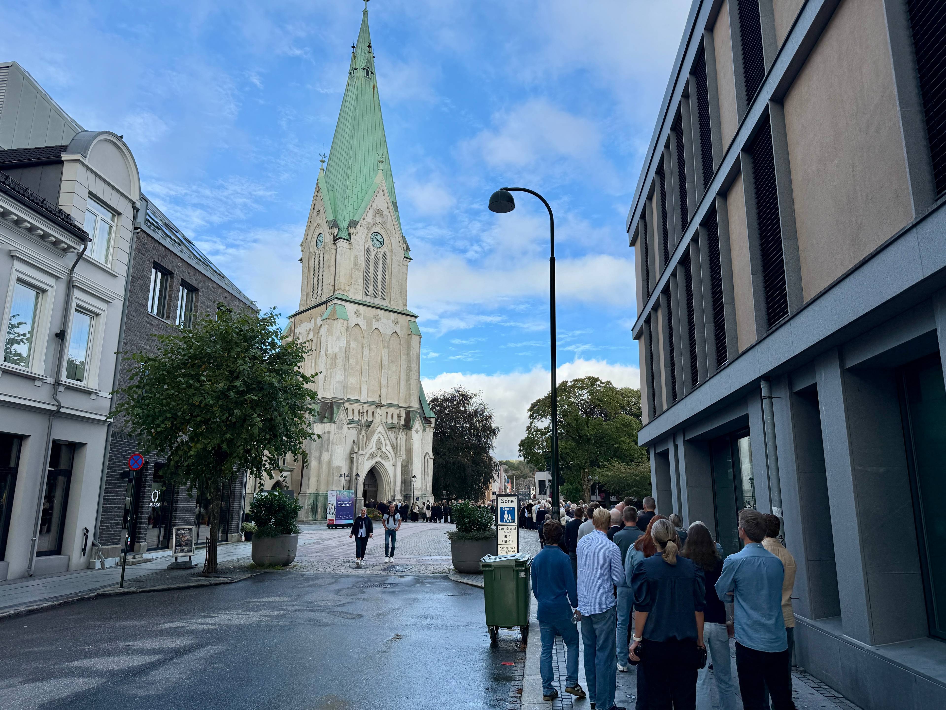 Mennesker står i kø på fortauet ved en kirke i en bygate.
