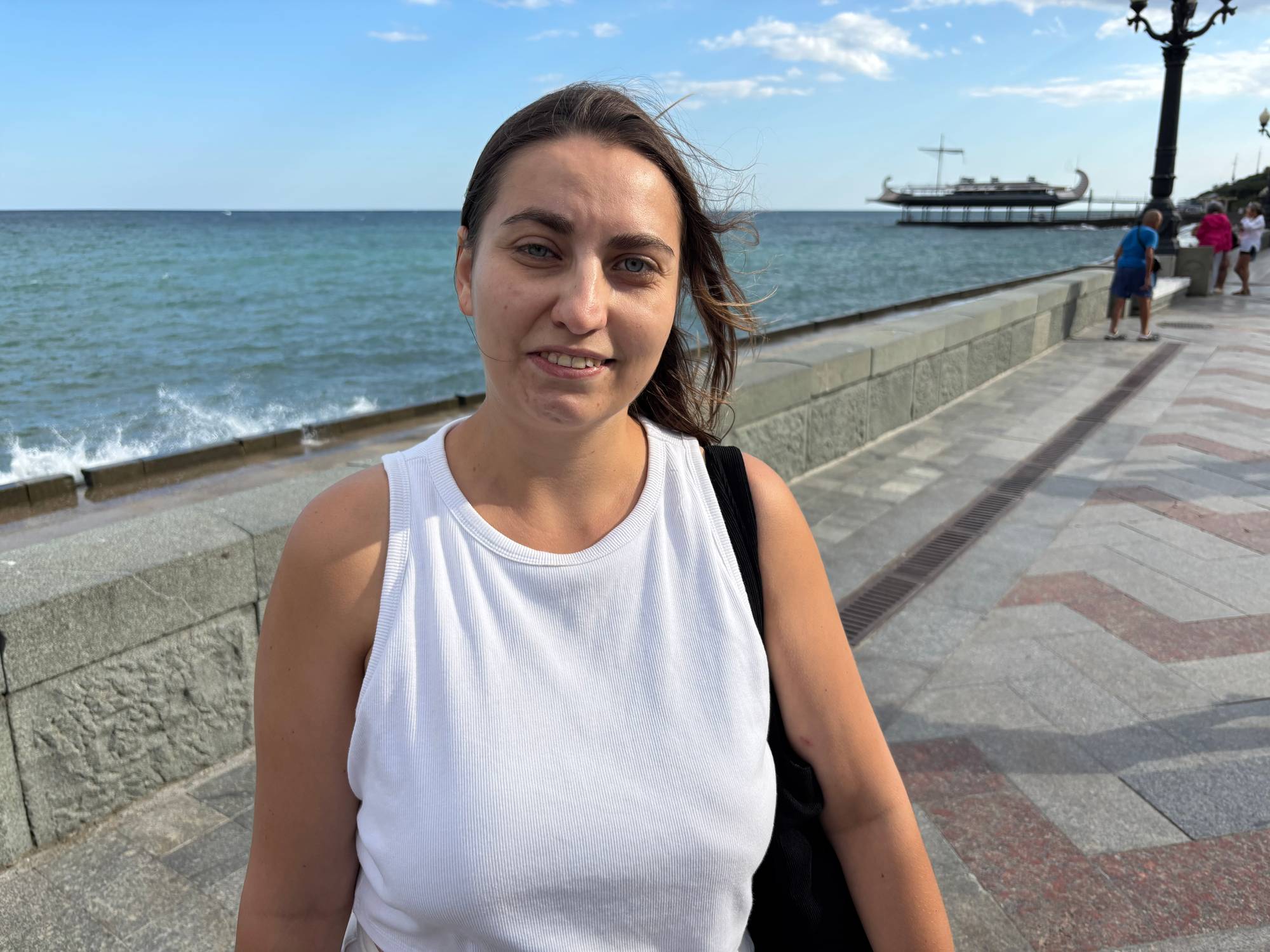 Kvinne i hvit t-skjorte ser i kamera. Hun står med ryggen mot havet på en strandpromenade.