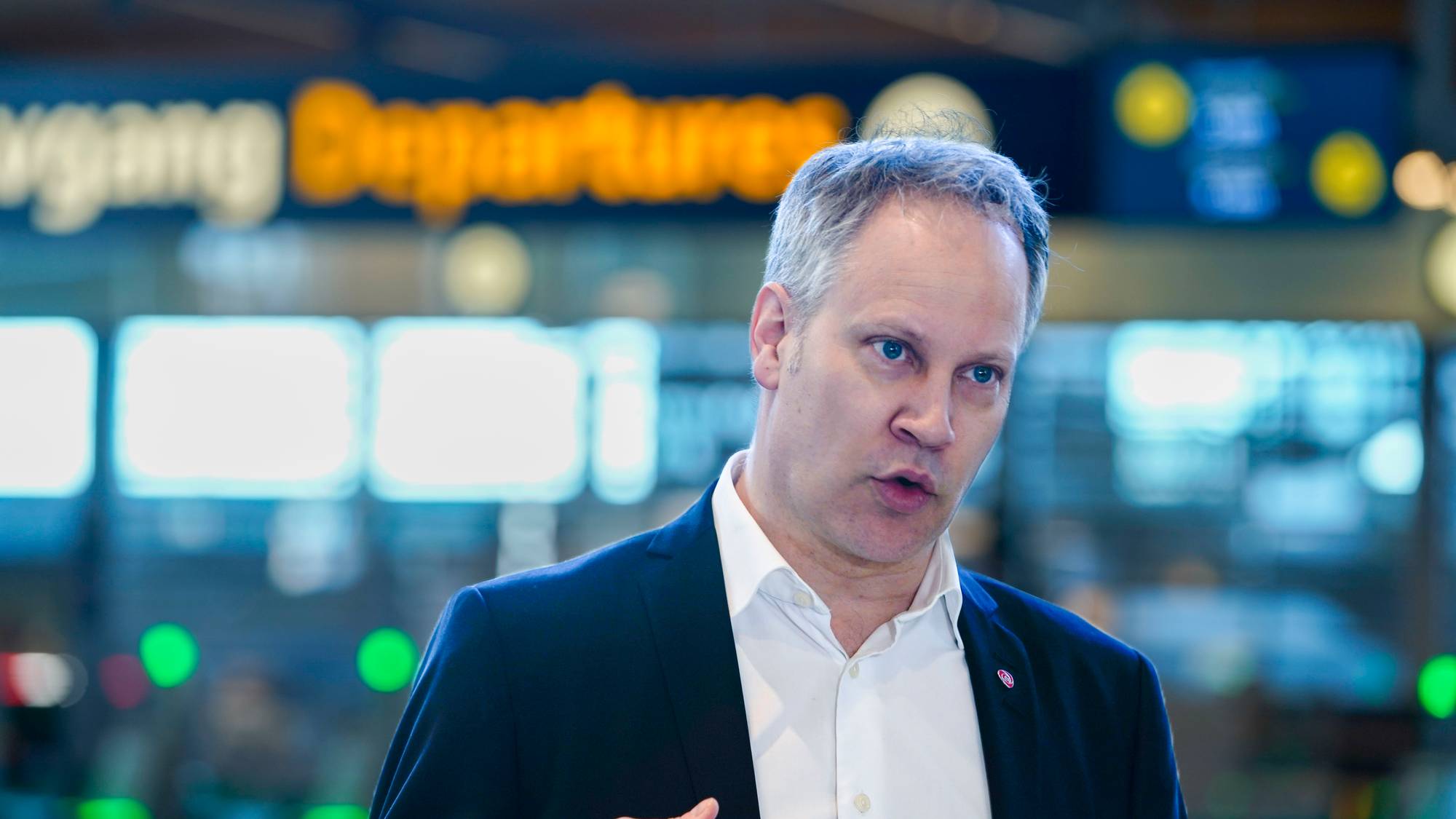 En person står i fokus og gestikulerer med hånden. I bakgrunnen er det en avgangstasjon med opplyste skilt. Omgivelsene virker travle, typisk for en flyplass. Det er høy belysning, og informasjonsskiltene viser avgangstider.