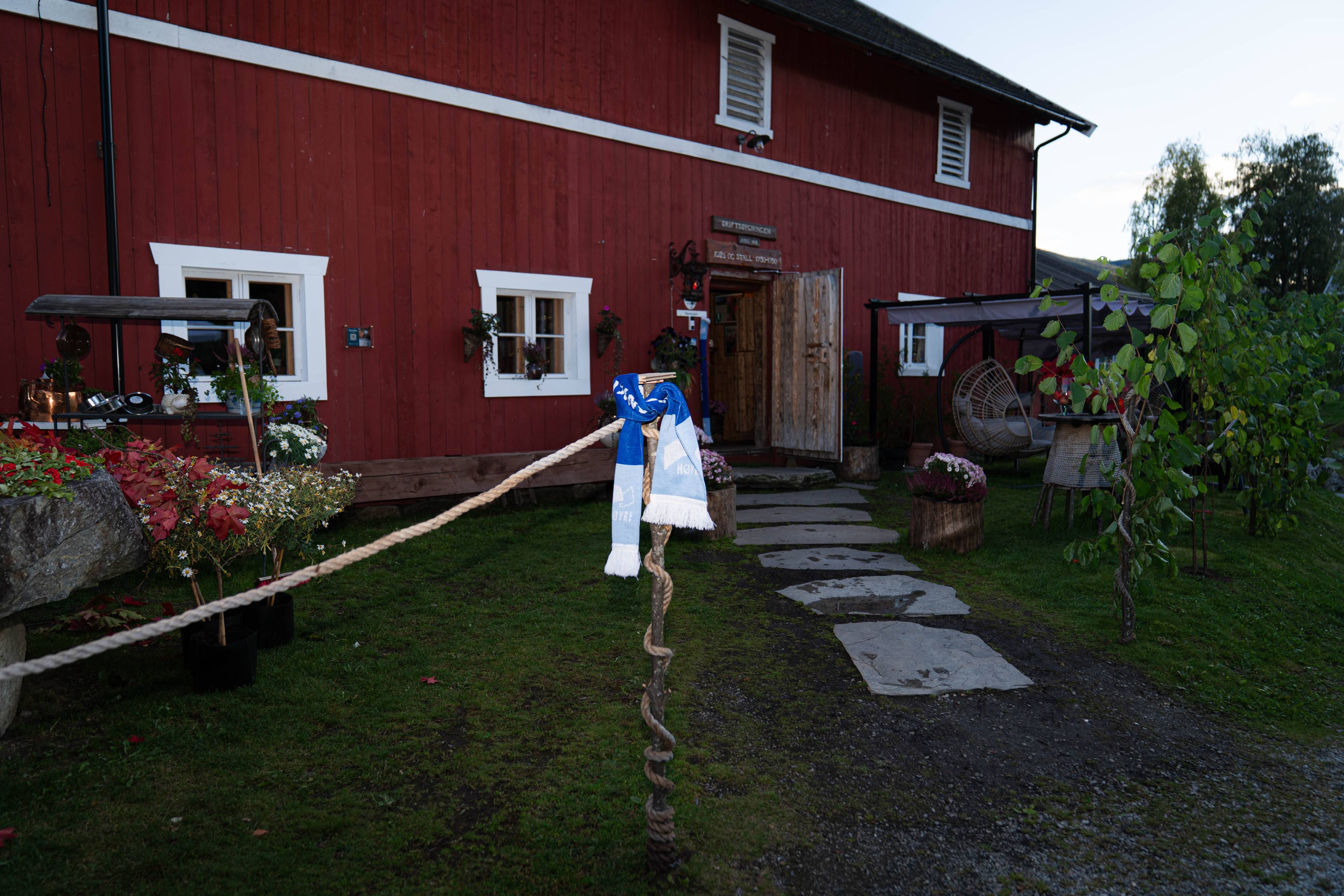 Et Høyre-skjerf henger på en stolpe foran en rød trebygning med en steinlagt sti til inngangen. (Bildebeskrivelsen er laget av en KI-tjeneste)