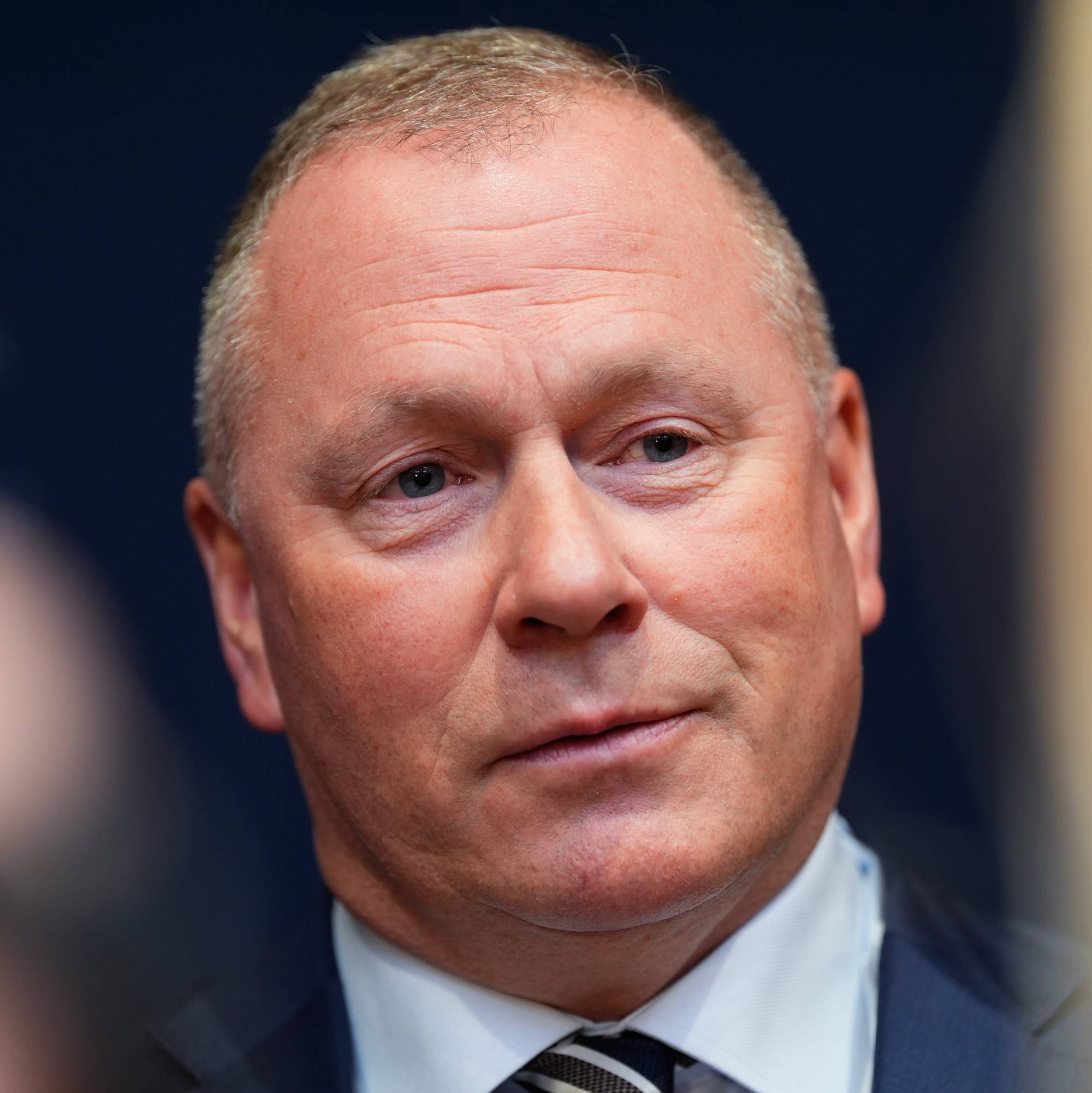 Bilde viser en mann i dress som deltar på en pressekonferanse. Han er omgitt av flere personer, som ser på ham mens han snakker. Bakgrunnen er uskarp, men antyder et profesjonelt miljø. Det er fokus på mannen i bildet, som ser ut til å være i en offentlig setting. (Bildebeskrivelsen er laget av en KI-tjeneste)