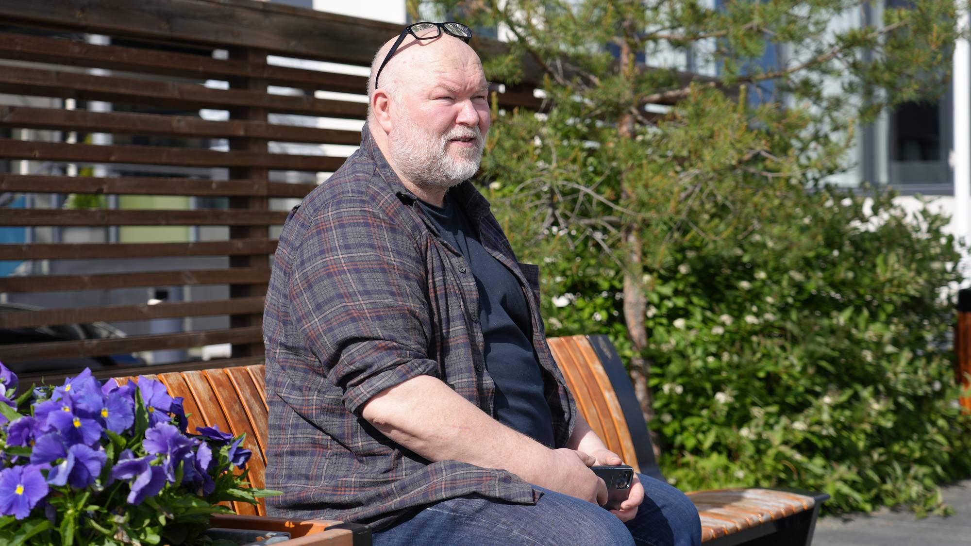 Stein Arne Rånes sitter på en benk med grønn plante bak seg og lilla blomster foran 