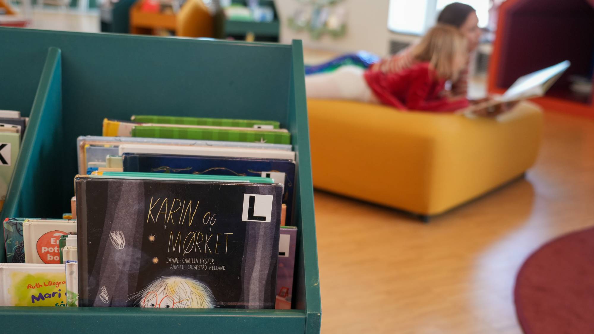 I forgrunn er flere små hyller med bøker i Stormen Bibliotek i Bodø. En mor og en datter ligger på en puff og leser på Stormen bibliotek i Bodø i bakgrunn.