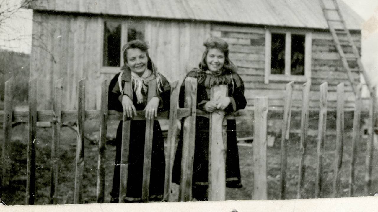 Bjørg og Tullik lener seg mot et gjerde foran skolestua, kledd i kofte