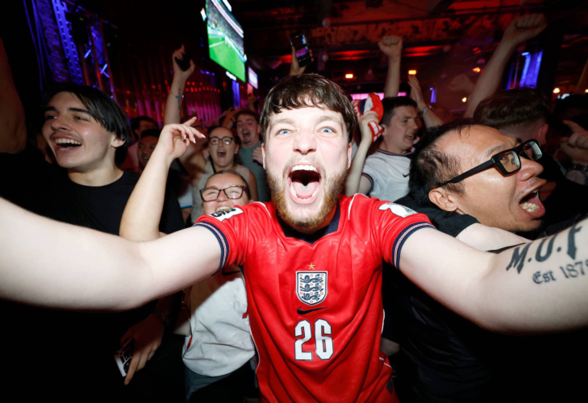Folk feirer i en pub mens de ser fotballkamp på skjerm. En mann i England-drakt jubler mot kamera. (Bildebeskrivelsen er laget av en KI-tjeneste)