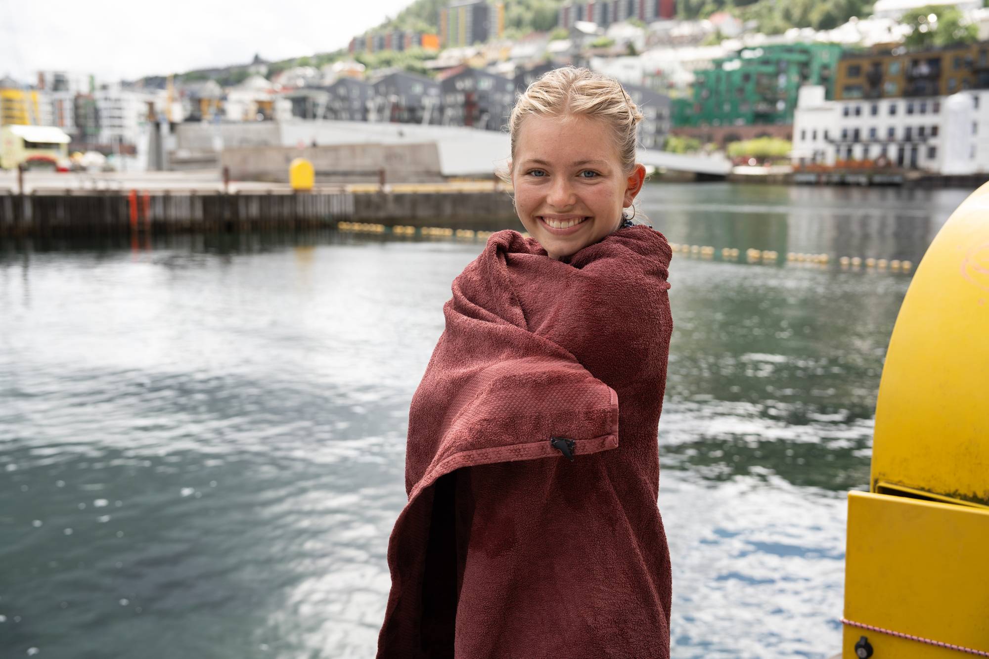 Smilende kvinne svøpt i et rødt håndkle ved en kai i Bergen med bybebyggelse i bakgrunnen. (Bildebeskrivelsen er laget av en KI-tjeneste)