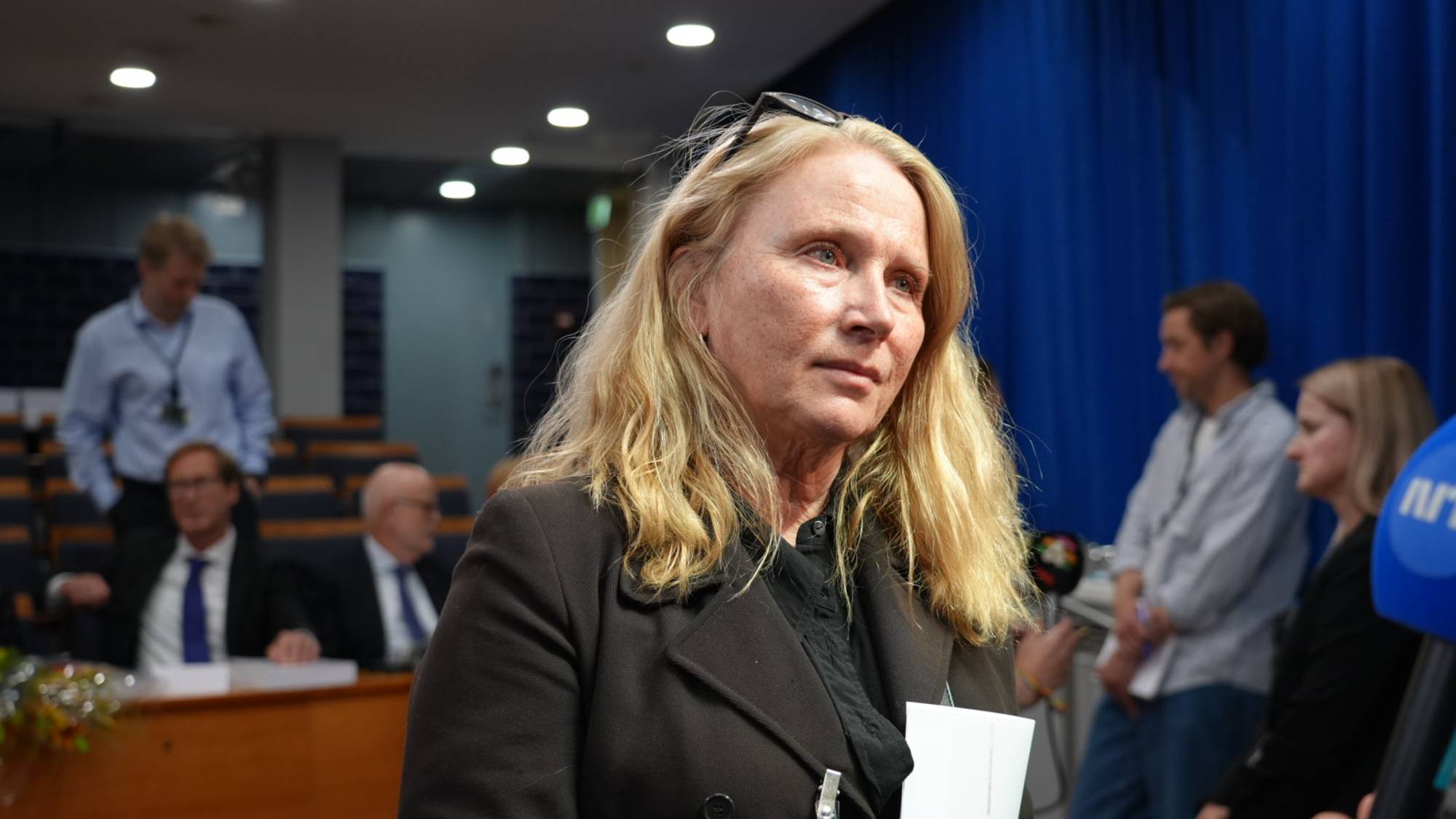 En kvinne med blondt hår står foran en blå bakgrunn. Hun har en mørk jakke på seg og et navnekort festet til jakken. Bak henne sitter menn i dress og en mann i lyseblå skjorte. Bildet er tatt innendørs, muligens i et konferanserom. (Bildebeskrivelsen er laget av en KI-tjeneste)