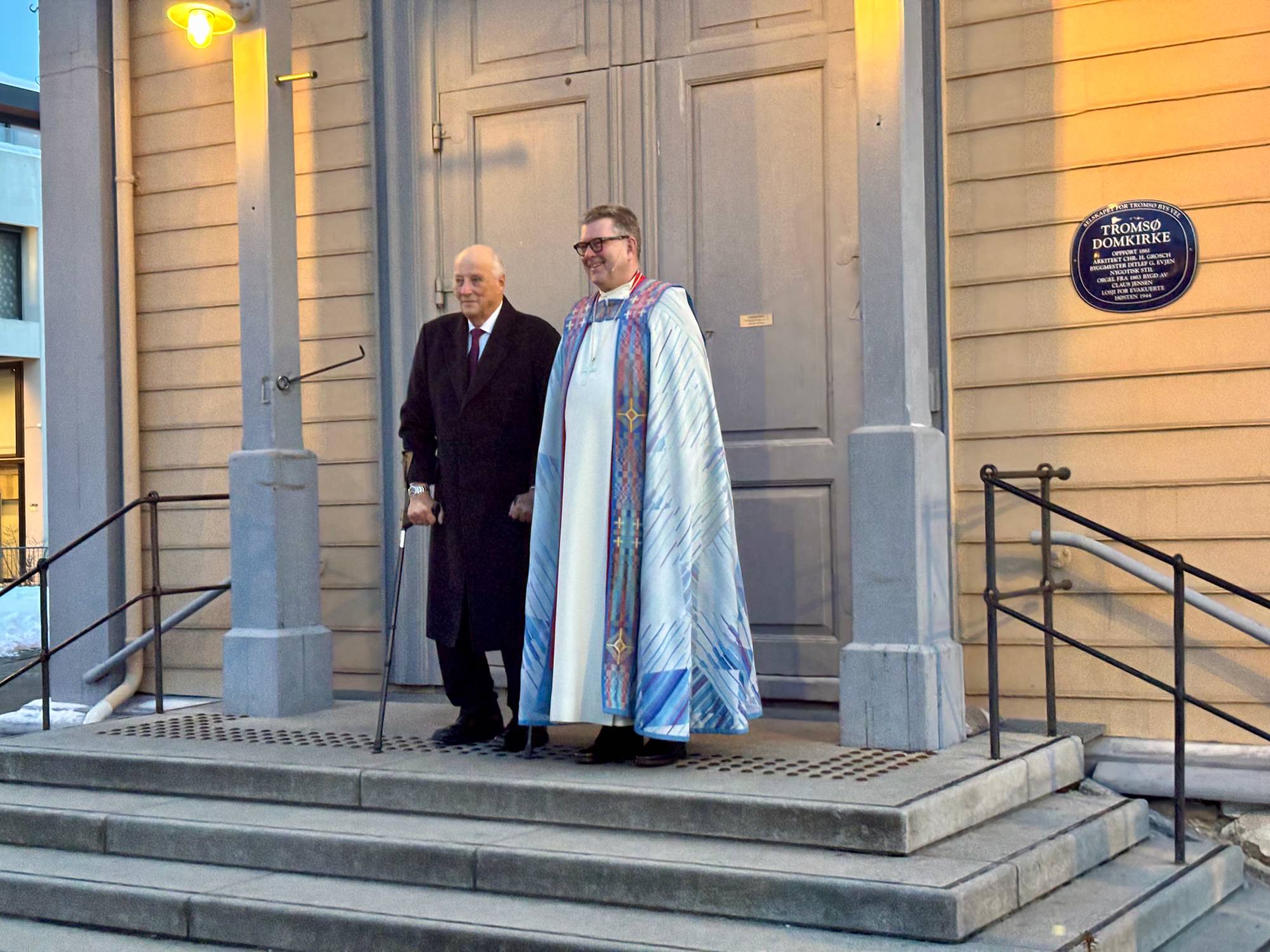En mann i sort frakk står foran en kirke med en stav, ved siden av en person i presteklær med fargerik kappe. De står på trappen til bygningen, som har en plakk med navnet "Tromsø Domkirke" synlig på veggen. Omgivelsene viser en vinterlig setting, med snø på bakken og dører som er stengt bak dem. (Bildebeskrivelsen er laget av en KI-tjeneste)